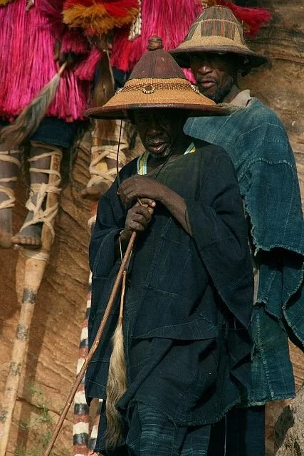 Two Dogon men wearing woven straw hats and dark indigo garments stand in front of a cliffside dwelling in Mali.