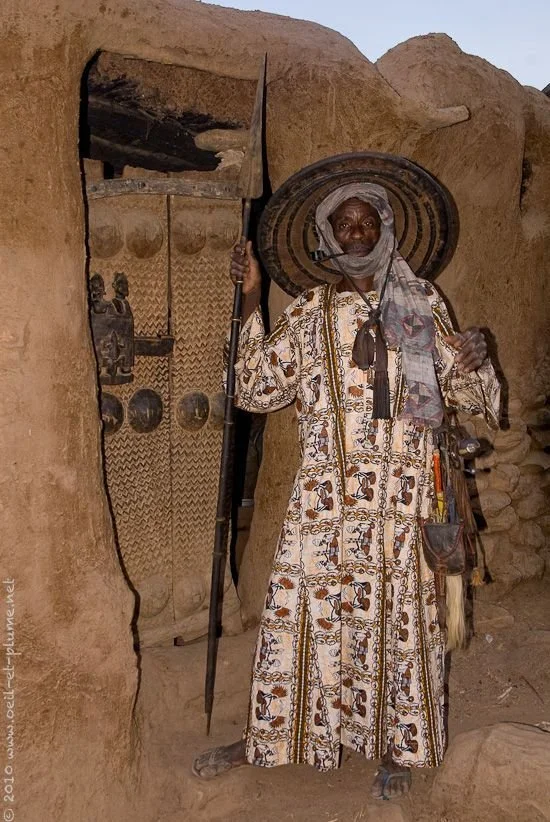 A Dogon man in a patterned robe and wide woven hat stands in the doorway of a mud-brick dwelling, holding a spear.