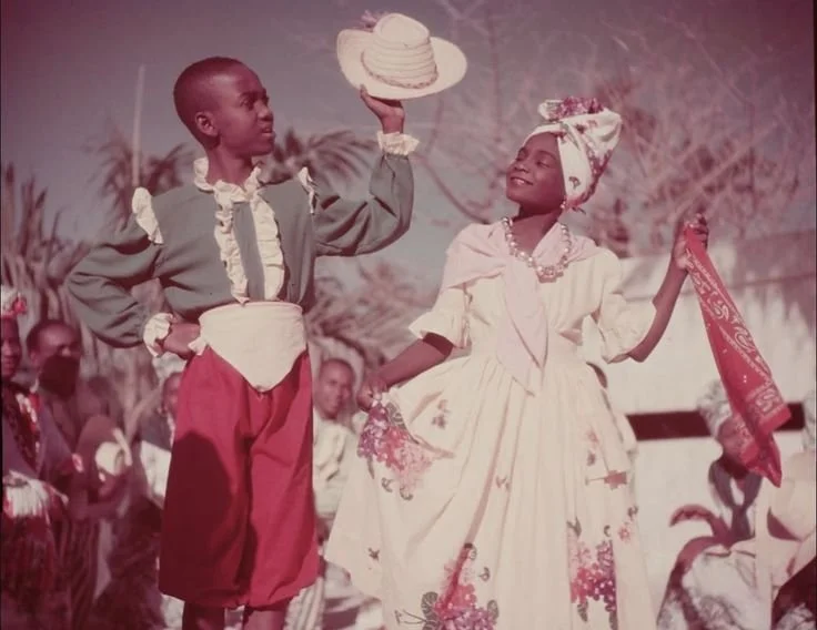 Haitian children performing a traditional dance in Port-au-Prince, wearing festive Creole attire including a madras headwrap, floral dress, and straw hat.