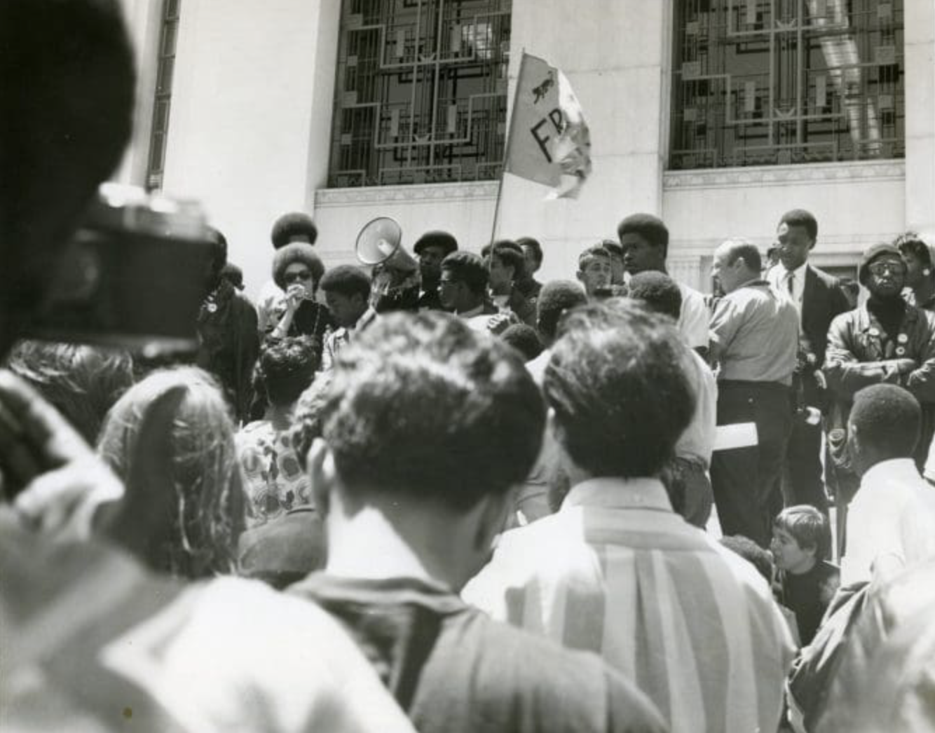 Photograph of a Black Panther Party rally with members addressing a crowd using a megaphone, with an FBP flag visible. Photographer unknown, c. late 1960s to early 1970s. Oakland Public Library Archives.