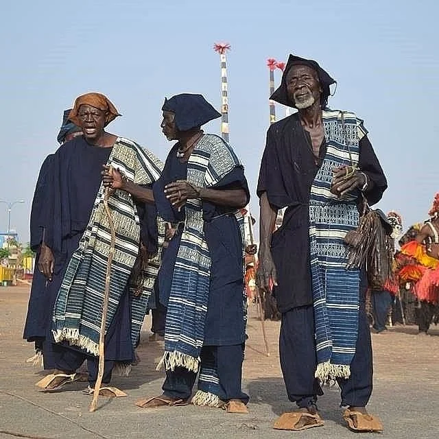 Three Dogon men wearing indigo garments and patterned woven shawls walk together.