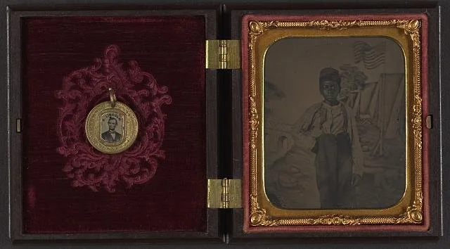 Unidentified African American boy standing in front of painted backdrop showing American flag and tents ; campaign button with portraits of Lincoln on one side and Johnson on the opposite side are attached to inside cover of case. Library of Congress