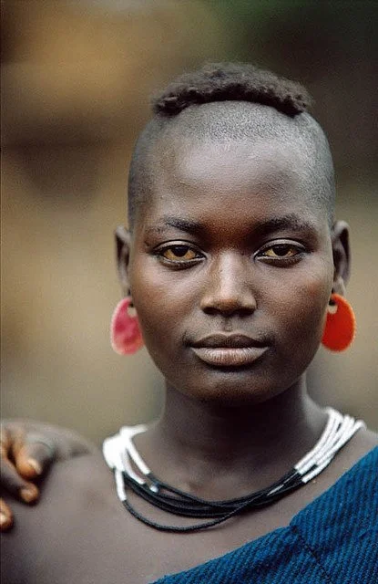 Suri woman from Ethiopia with a partially shaved head featuring a small patch of hair on top, a sign of feminine beauty. Sourced from Kwekudee.