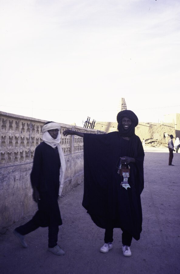 Two Wodaabe men stand outside in front of a perforated wall. 