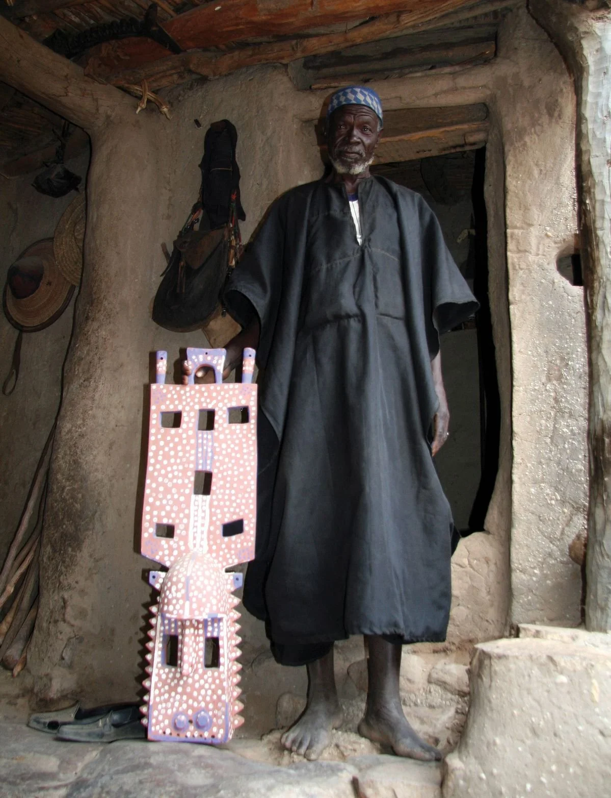 A Dogon man stands at the entrance of a mud-brick dwelling in Mali, holding a painted kanaga mask.
