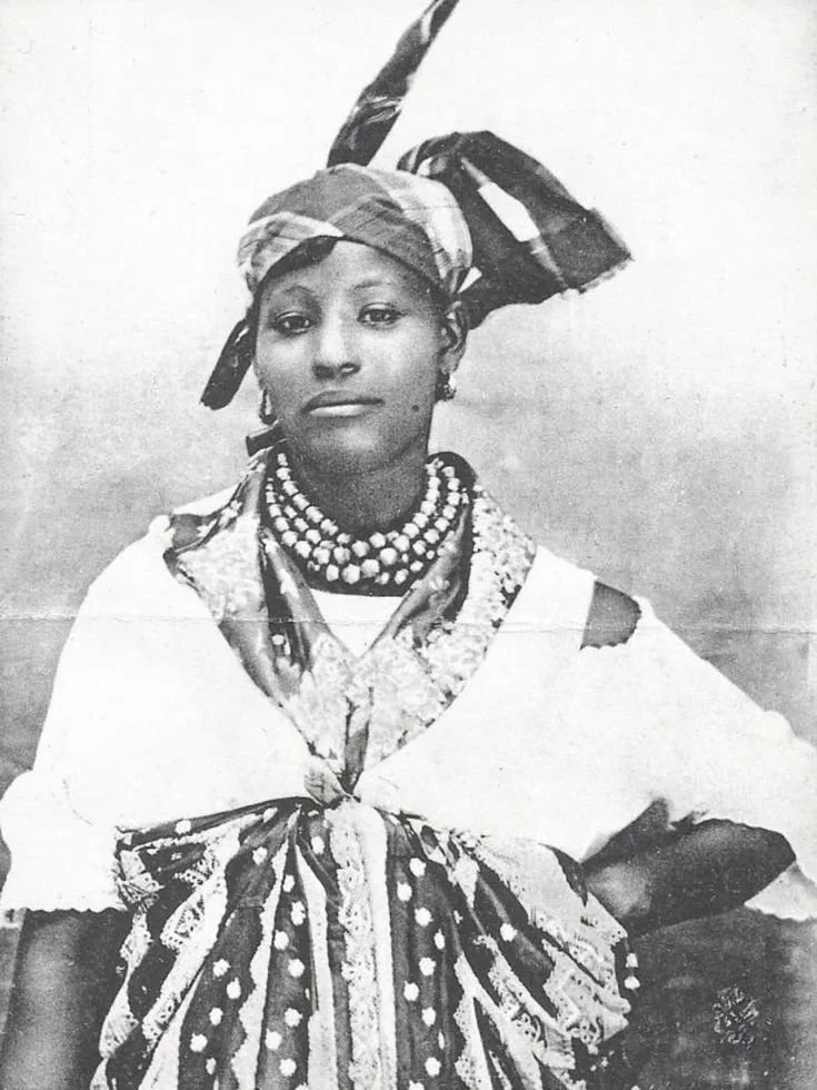 Caribbean woman wearing a karabela (quadrille) dress, adorned with layered bead necklaces and a madras headwrap