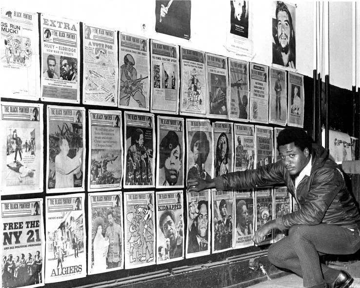 Photograph of Craig C. Williams adjusting a news cover featuring Malcolm X on a wall of Black Panther newspapers at 1928 Columbia Avenue in Philadelphia. Taken on October 23, 1969 by photographer Jay Beachem.