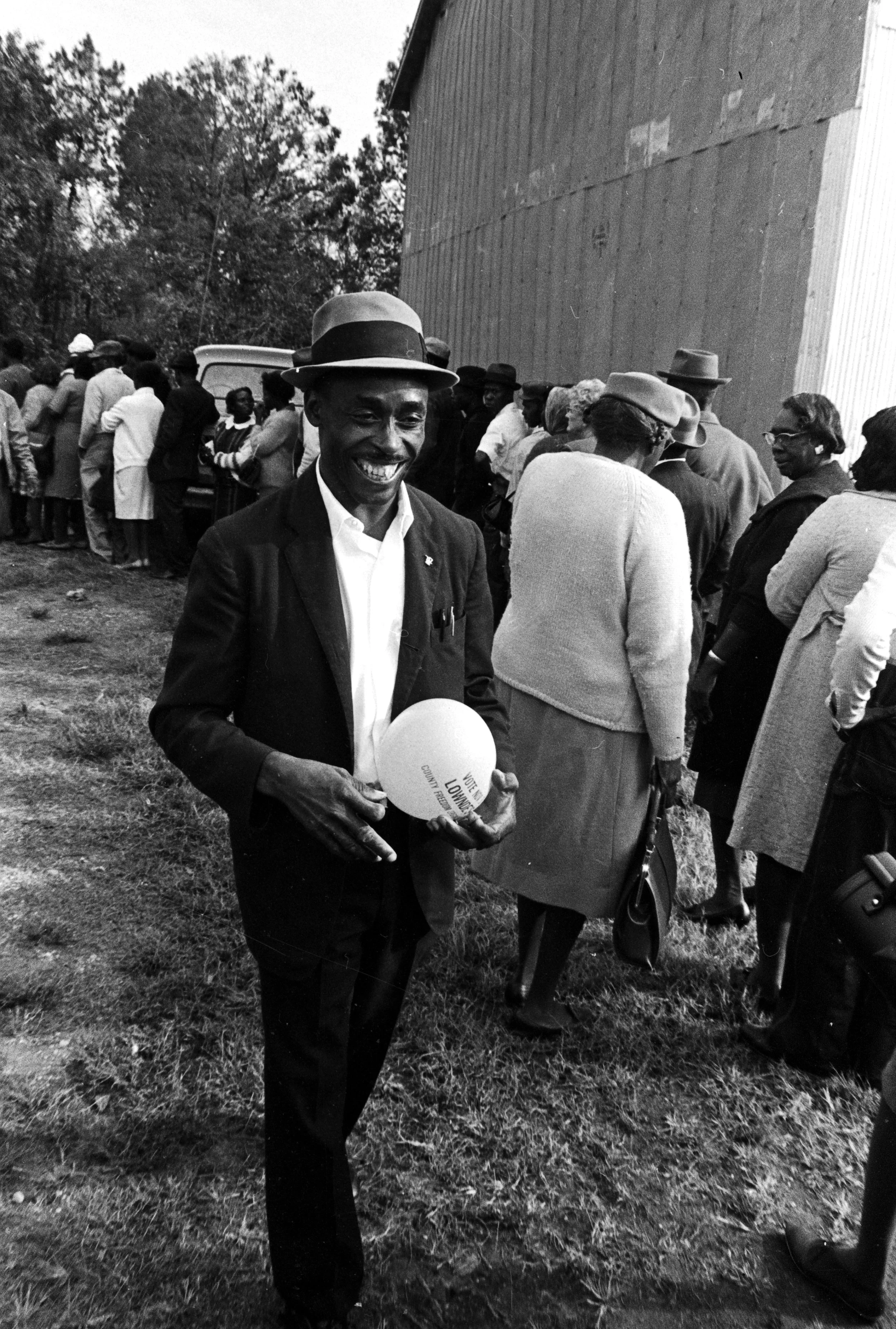 John Hulett walking past people standing in line to vote in Lowndes County, Alabama in 1966. He is holding a balloon that reads, “Vote Nov. 8 Lowndes County Freedom Organization.” © Alabama Department of Archives and History, Jim Peppler Southern Cou