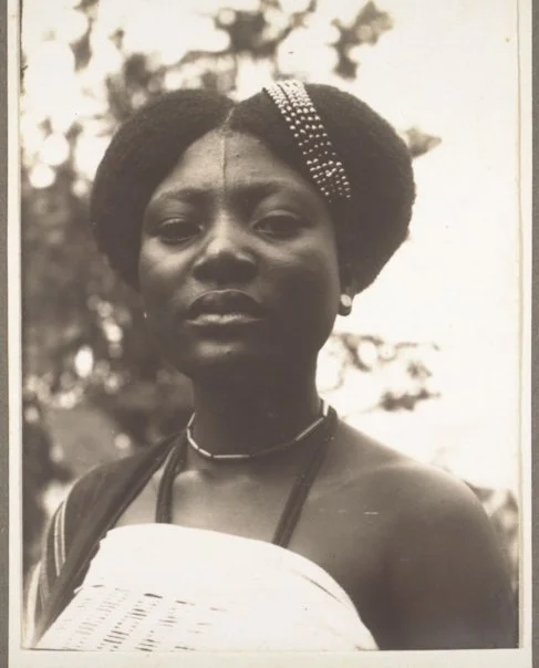 Funmilayo Ransome-Kuti photographed with her hair parted down the center, swept into a high, sculptural updo and adorned on one side with a beaded hairpiece. Sourced from Eccentric Yoruba. 