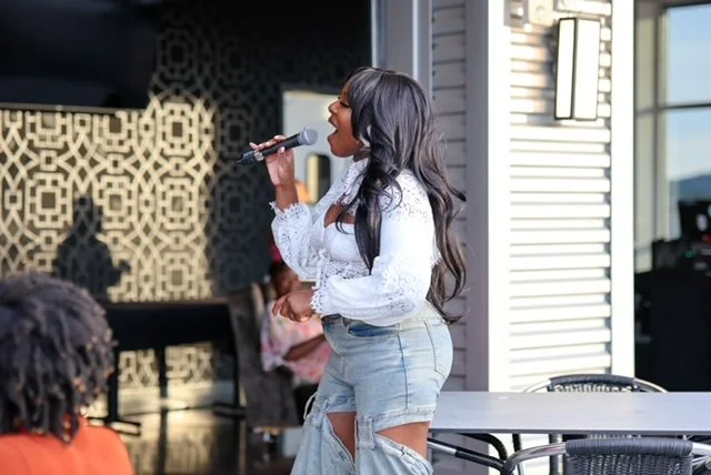 A woman with long dark hair singing into a microphone indoors, wearing a white blouse and light blue ripped jeans.