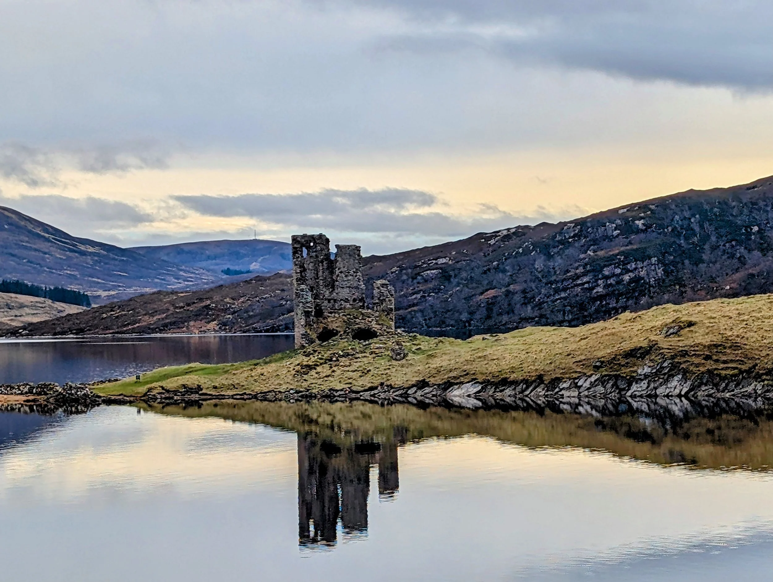A Scottish Highland loch with ancient stone tower ruins reflected in still water, and mountains in the background — the landscape that inspires Lovely Mama