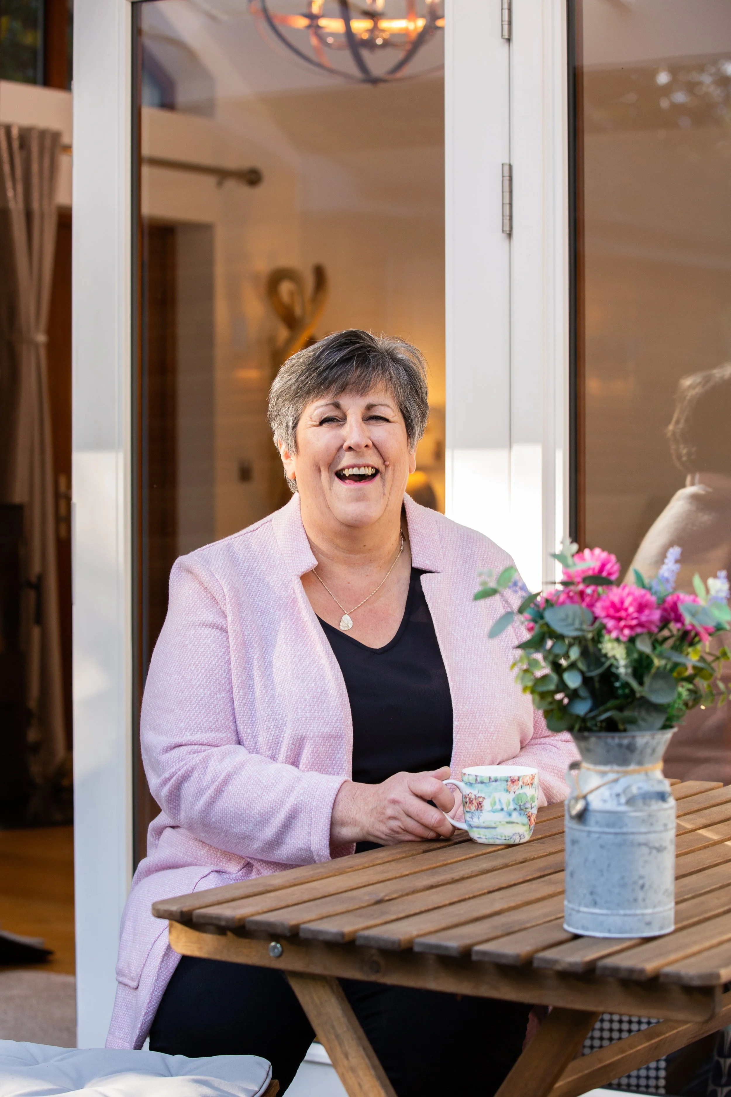 Shona, founder of Lovely Mama, smiling at an outdoor table surrounded by pink flowers