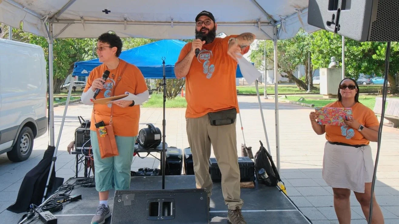 Three people standing under a tent, with two holding microphones and one holding a colorful box. They are wearing matching orange shirts and appear to be at an outdoor event. Equipment and a speaker are visible in the background.
