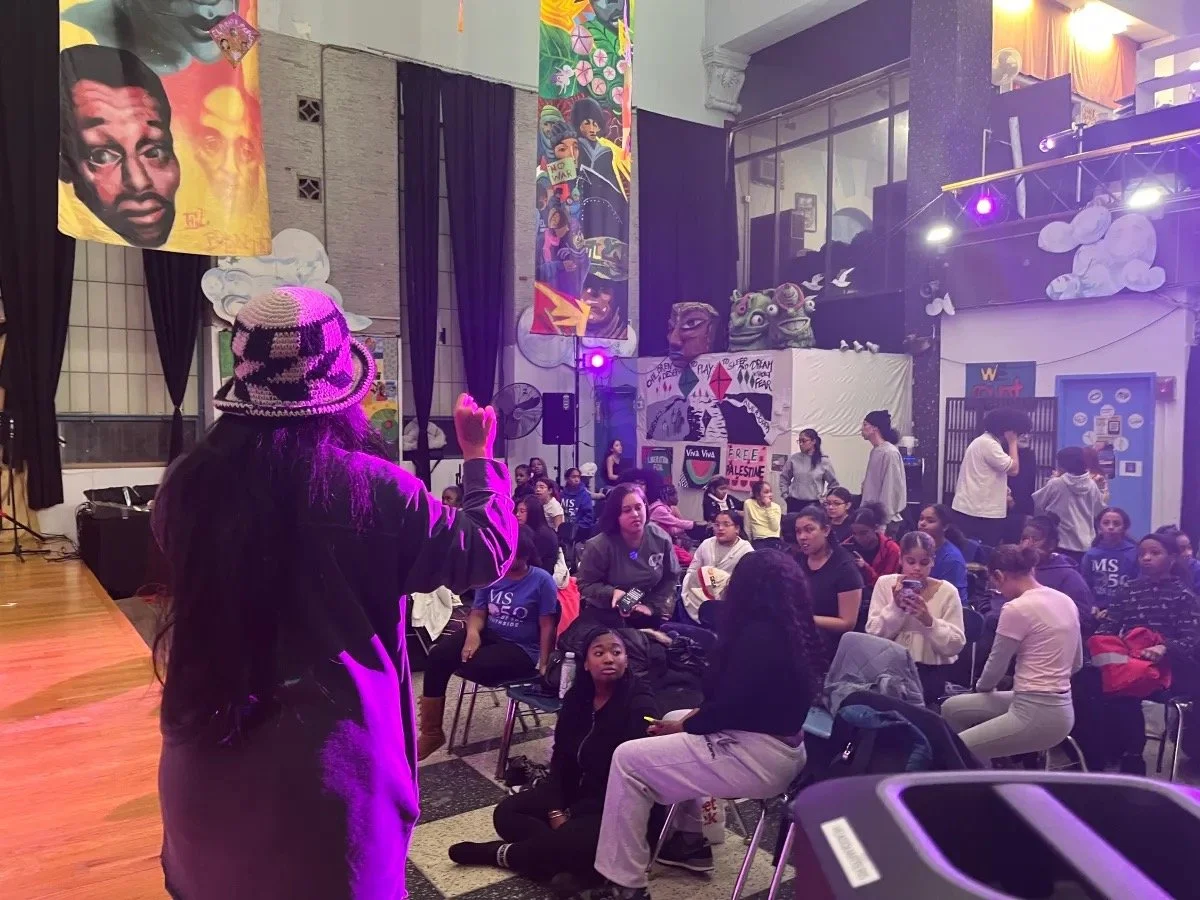 A person facing an audience in an indoor venue, with colorful art and banners hanging from the ceiling. The audience is seated, watching attentively.