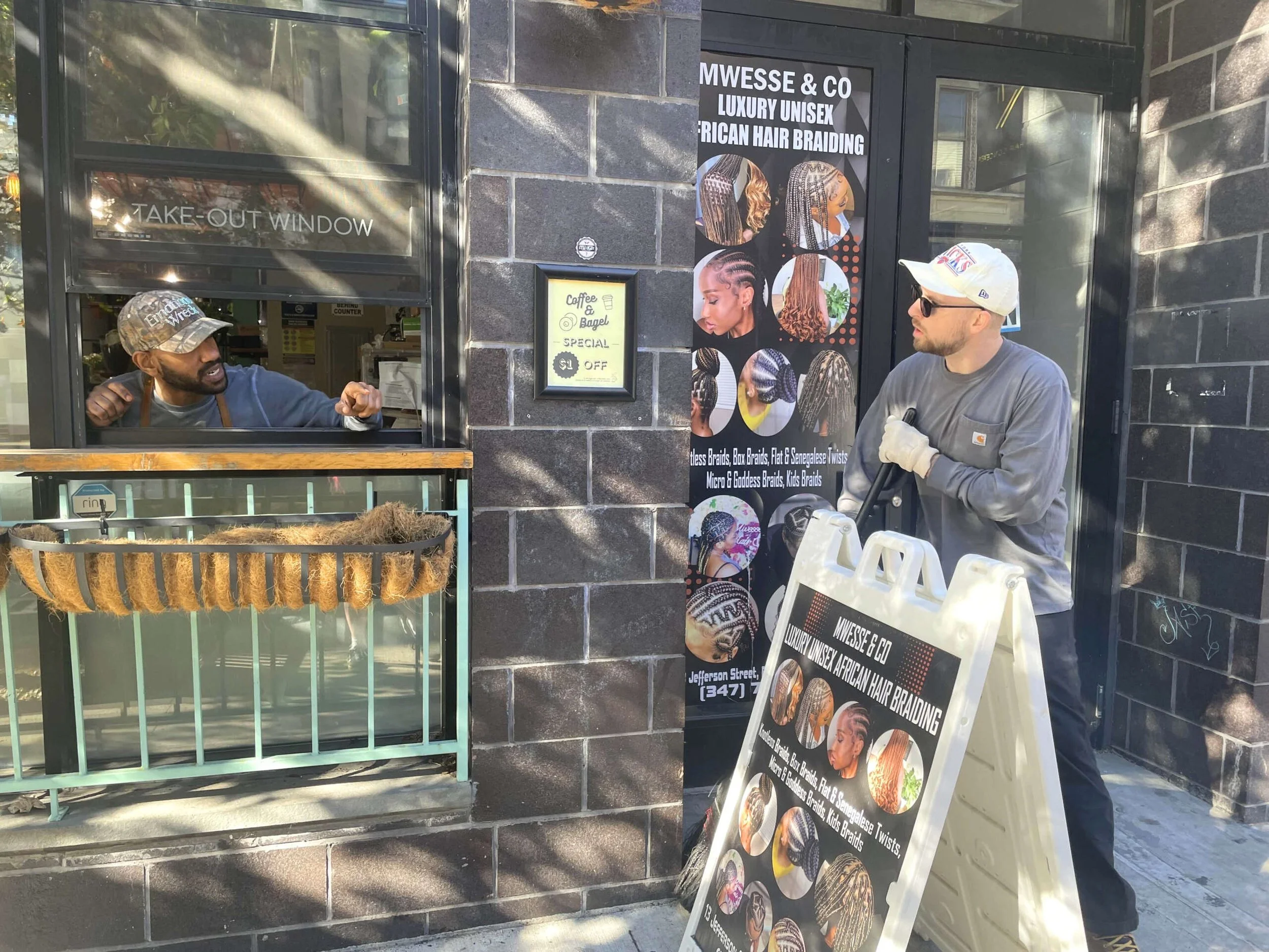 Two men interacting at a take-out window next to a storefront promoting luxury unisex African hair braiding, with signs displaying coffee and bagel specials.