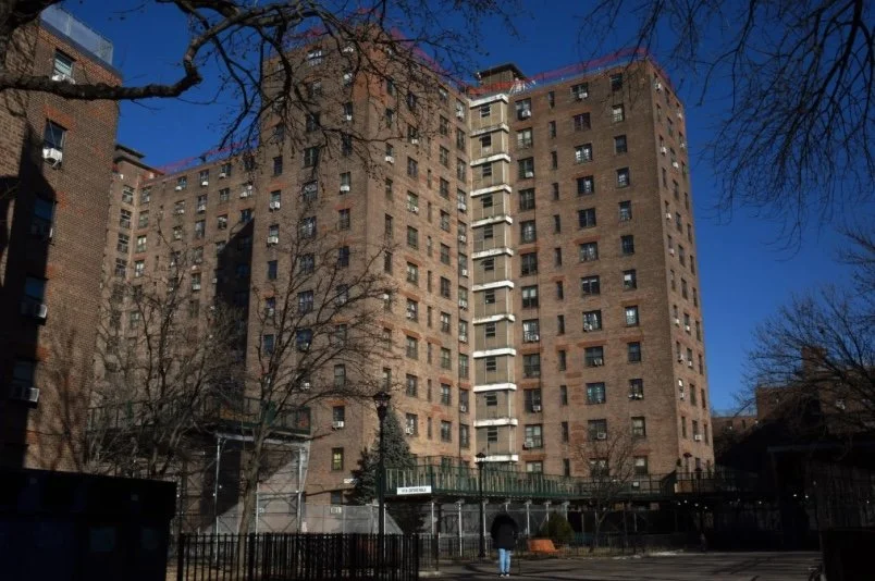 Large brick apartment building with multiple floors, trees and a walkway in the foreground, set against a clear blue sky.