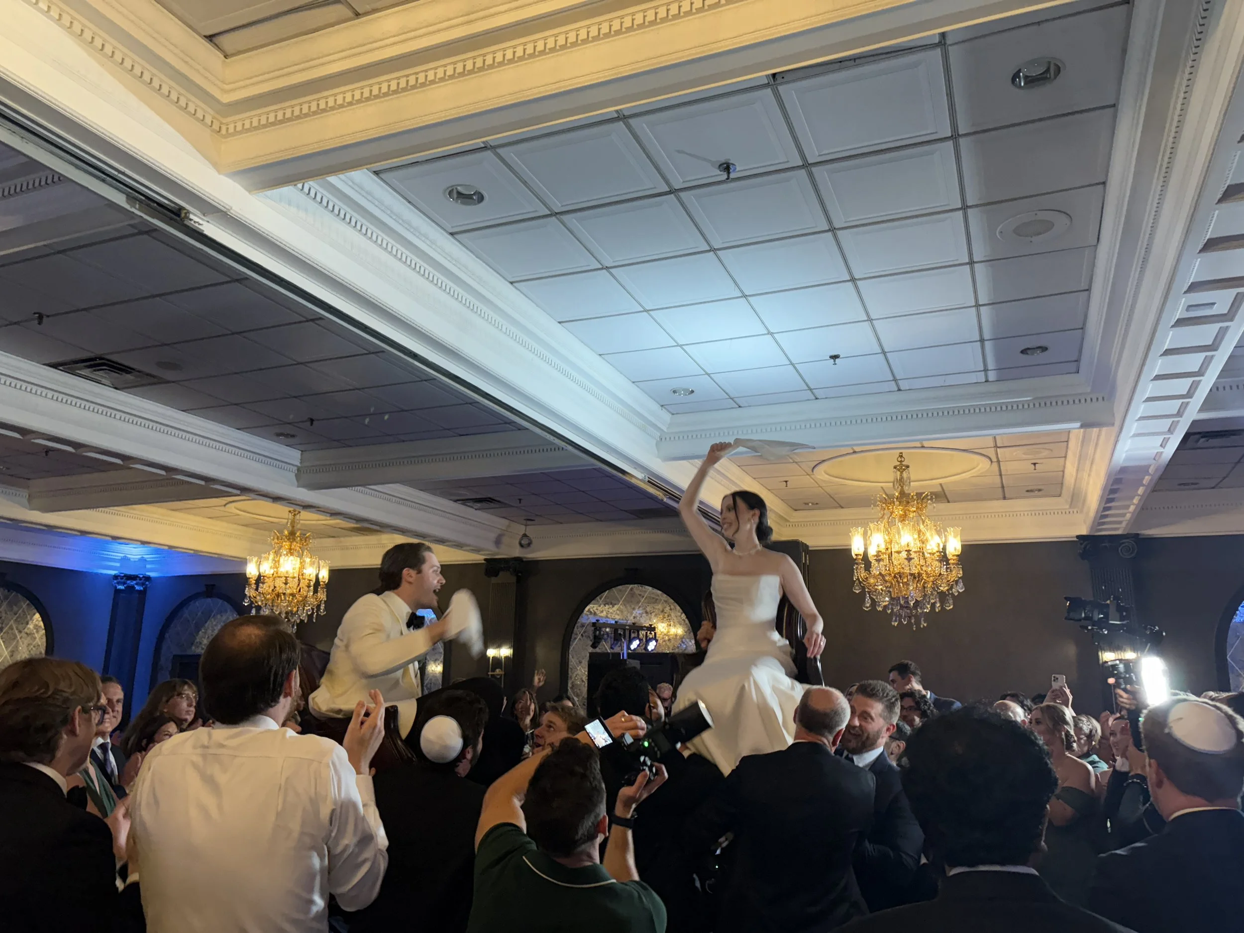 Wedding celebration with a bride and groom dancing on a chair, surrounded by guests in formal attire at an elegant hall with chandeliers and decorative ceiling.