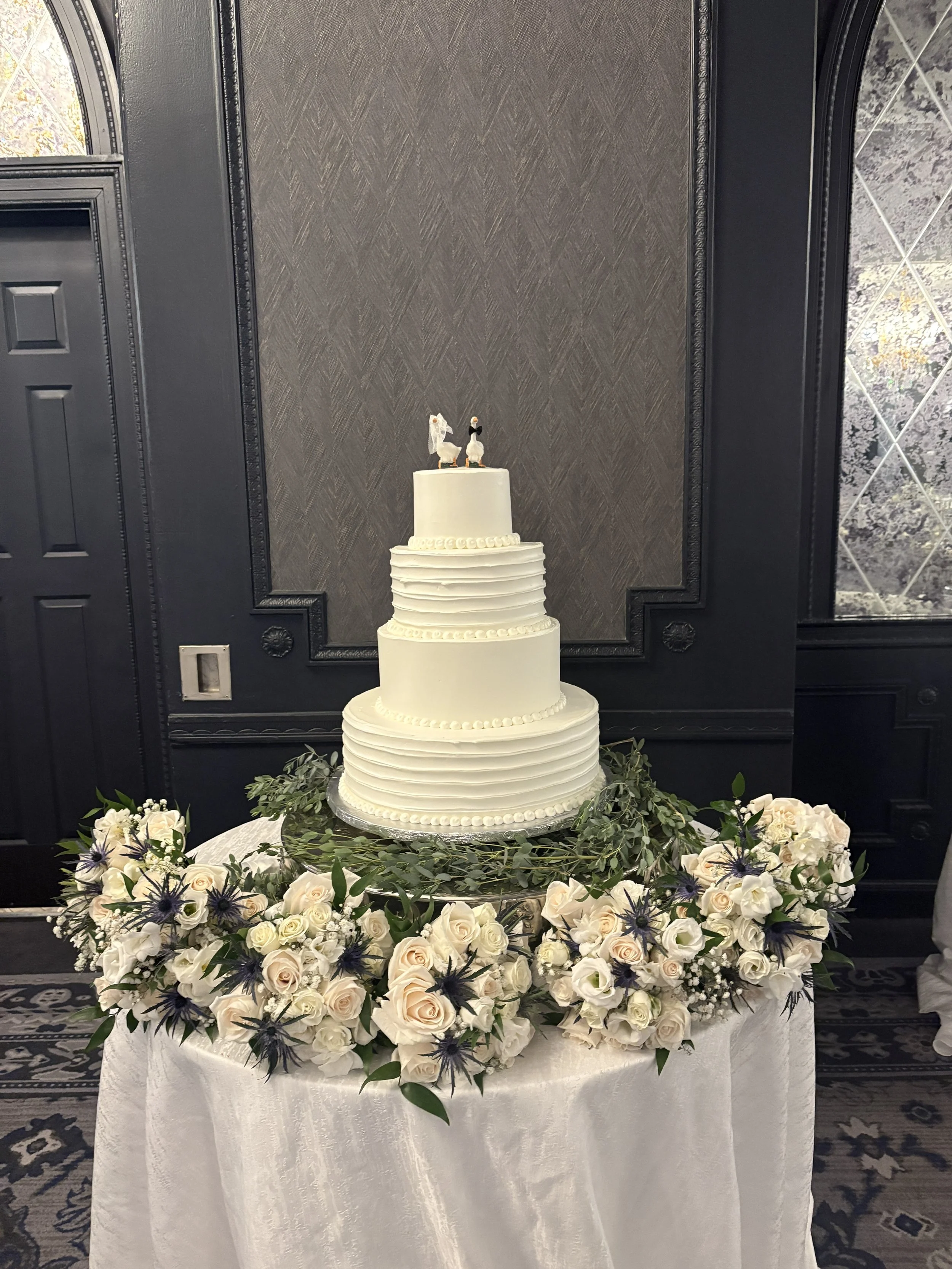 A tiered white wedding cake with a bride and groom topper, surrounded by white roses and greenery on a round table with a white cloth. The background features a dark ornate wall with textured patterns and a nearby window with decorative glass.