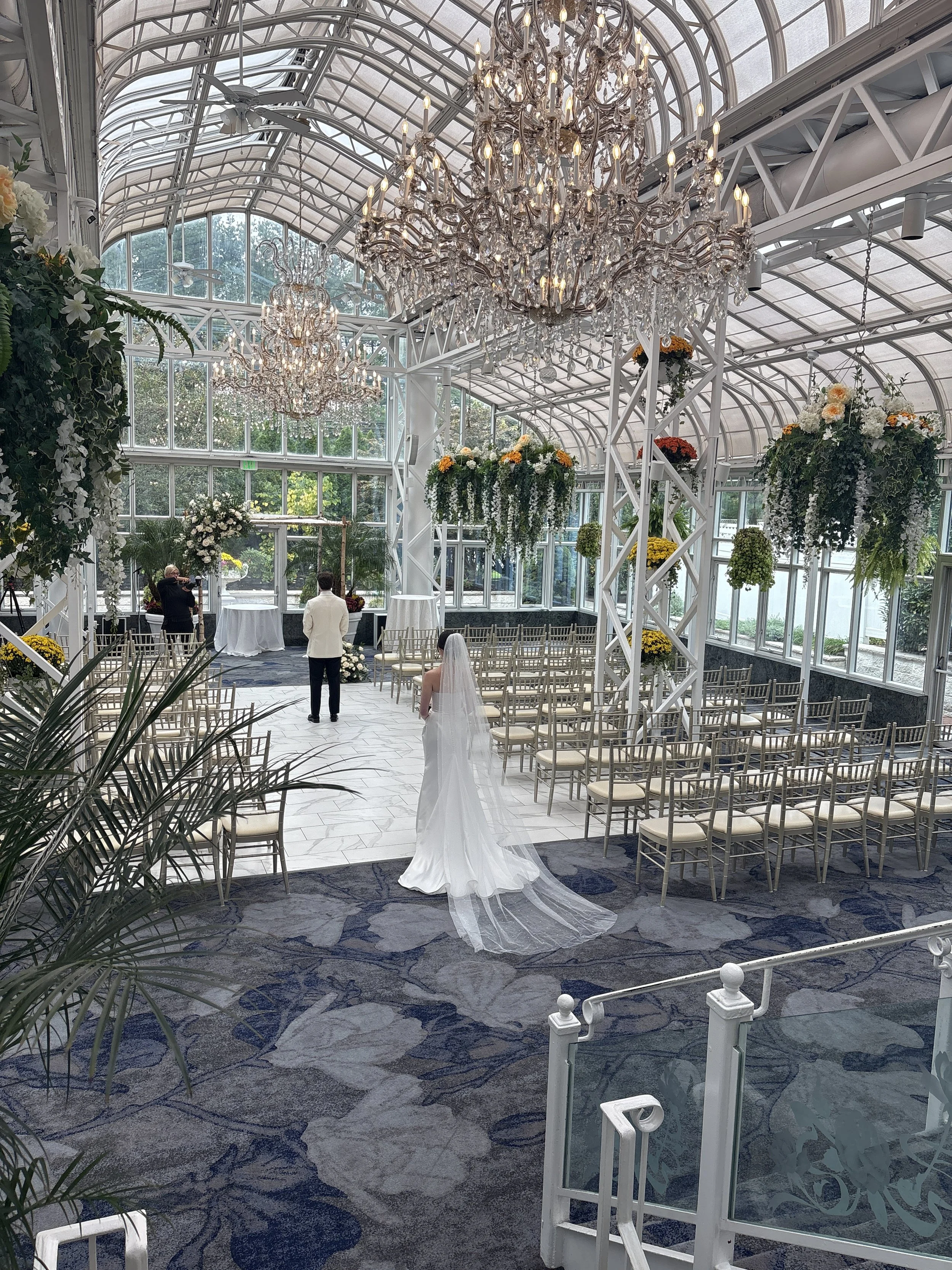 A bride in a white wedding dress and veil standing in a glass conservatory with chandeliers and floral decorations, preparing for a wedding ceremony.