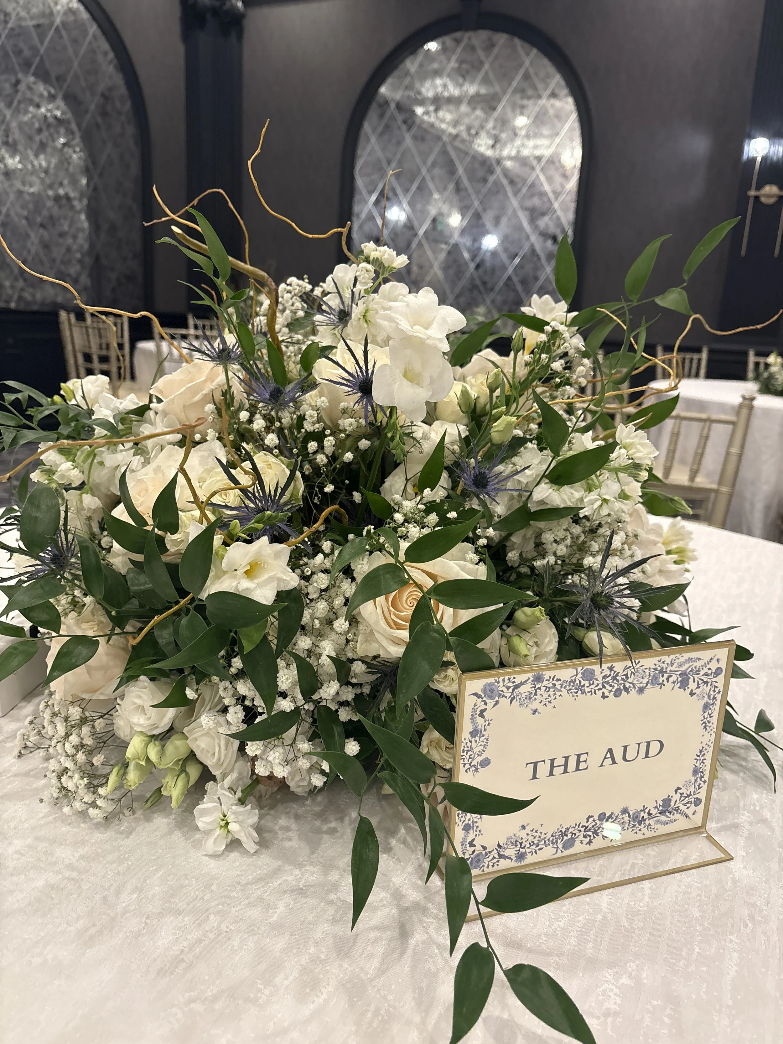 Elegant flower centerpiece with white roses, hydrangeas, baby's breath, and purple thistles, on a table with a sign reading 'The AUD'.