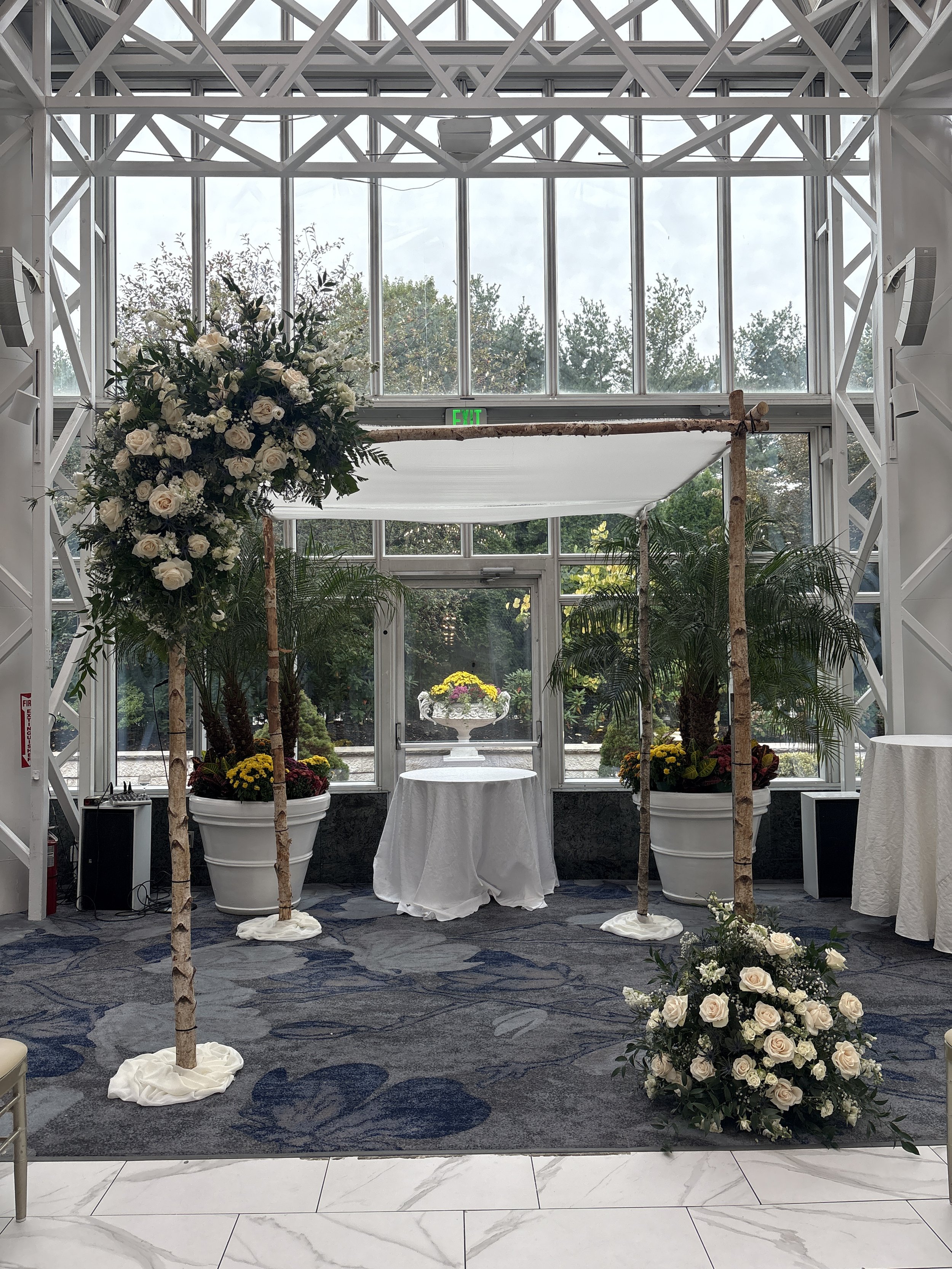 Wedding ceremony setup with a floral arrangement arch and potted palms inside a glass-enclosed building.