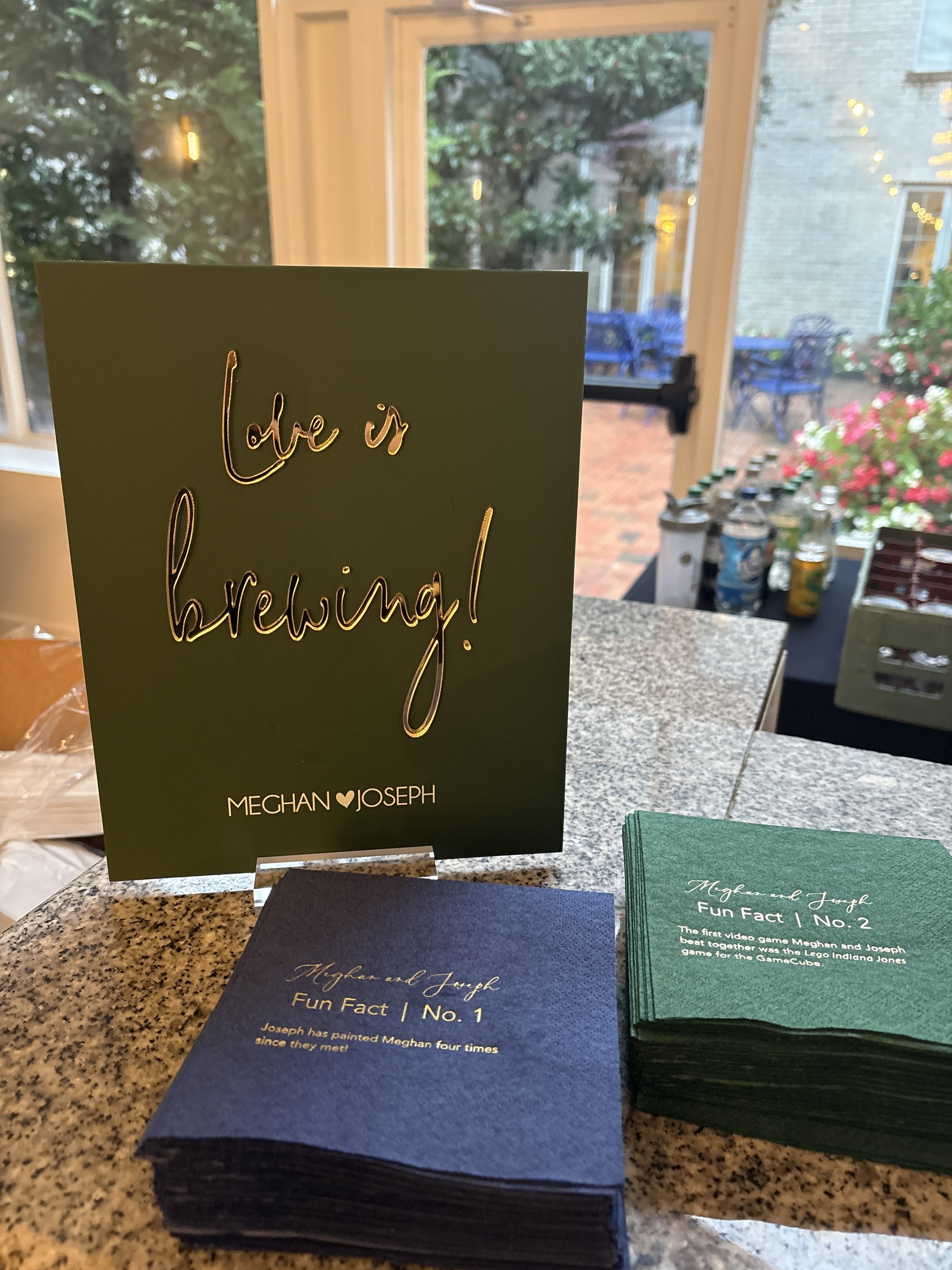 A decorative sign with gold lettering reads 'Love is brewing!' with the names Meghan and Joseph beneath it. There are stacks of cards nearby with fun facts about Meghan and Joseph, placed on a granite countertop. In the background, through a window, there is an outdoor patio with chairs, tables, and potted plants.