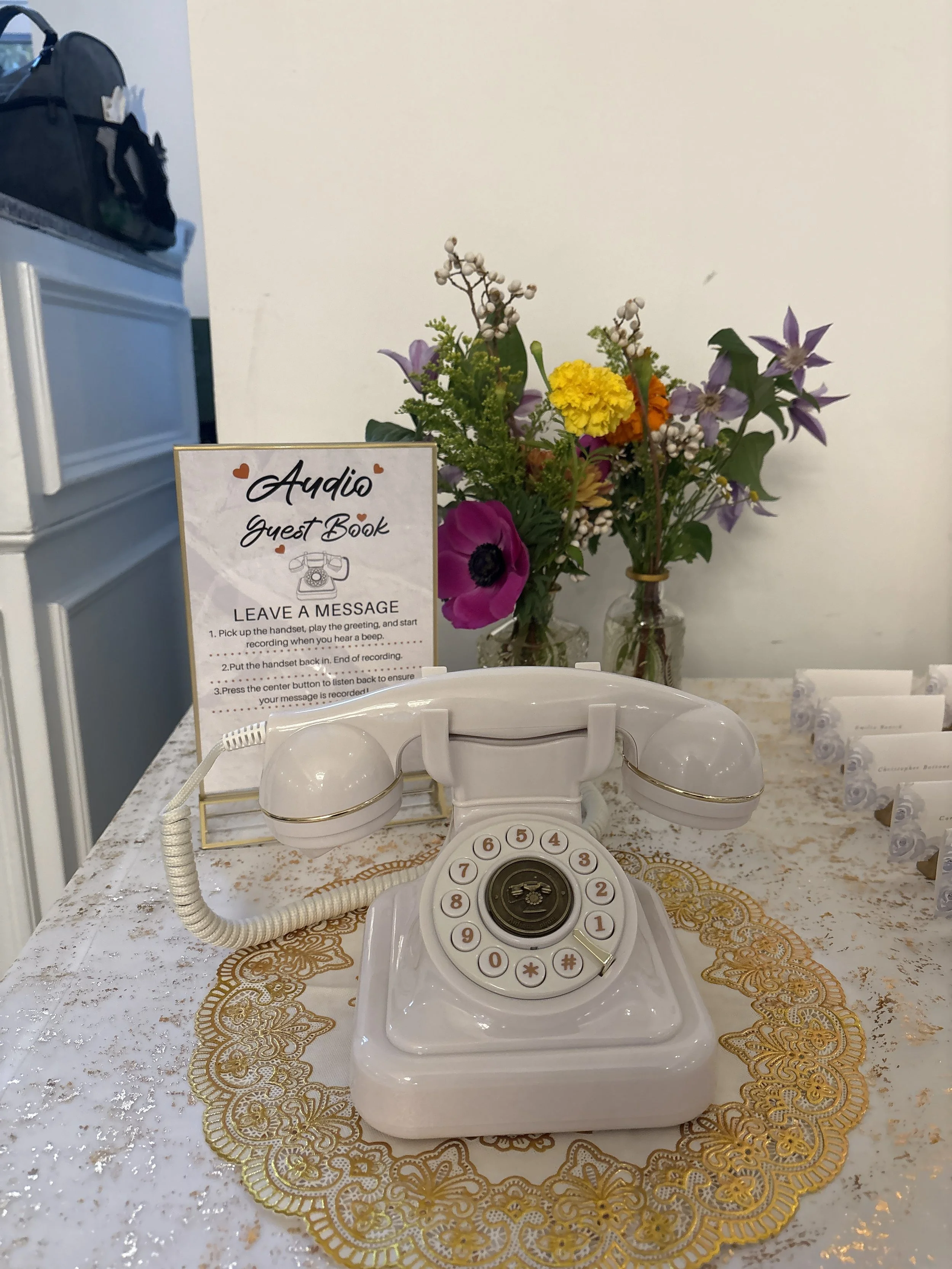 Vintage white rotary dial telephone on a decorative tablecloth with a gold lace design, next to a sign for an audio guestbook and a small bouquet of colorful flowers in vases.