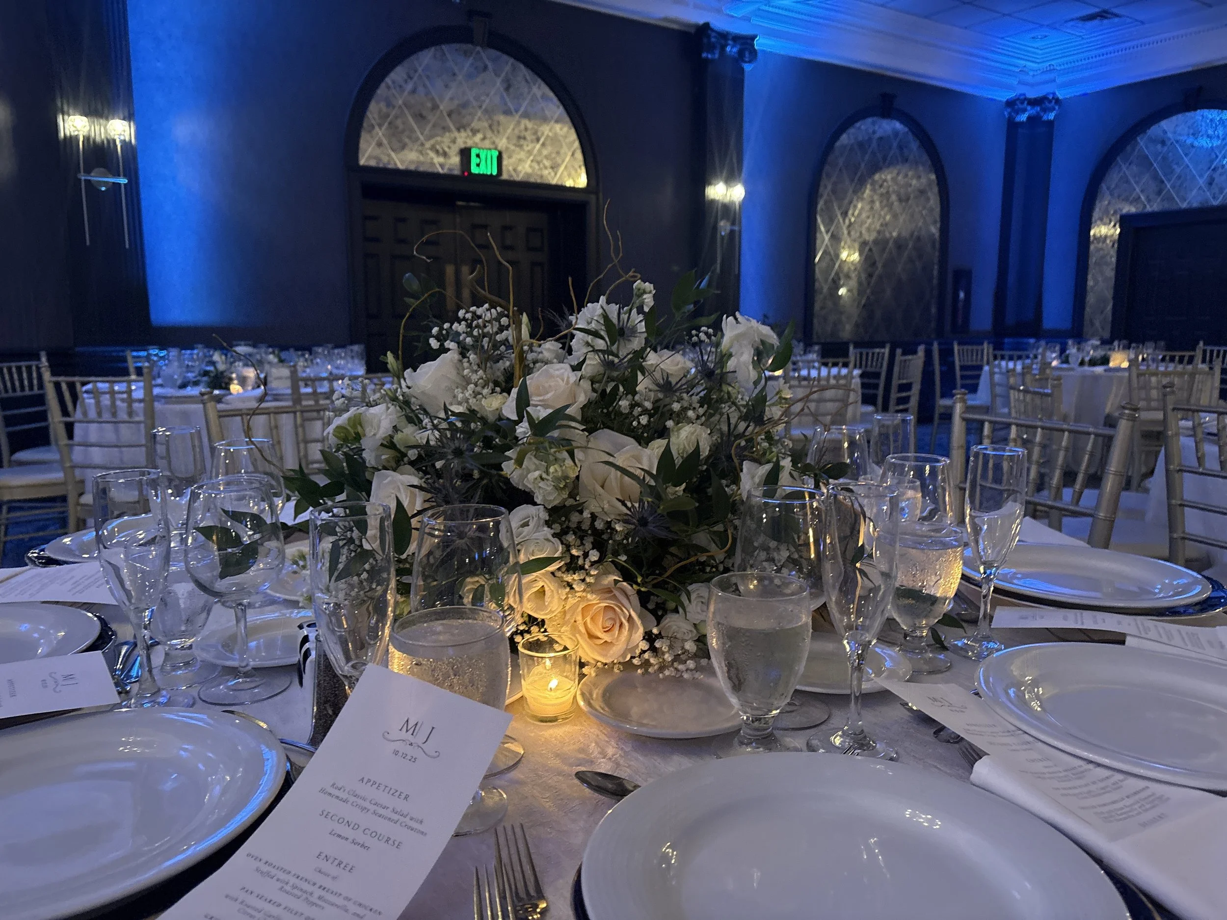 Elegant banquet table set for a wedding reception with white floral centerpiece, multiple glasses, plates, silverware, and a printed menu, in a dimly lit room with blue lighting and arched windows.