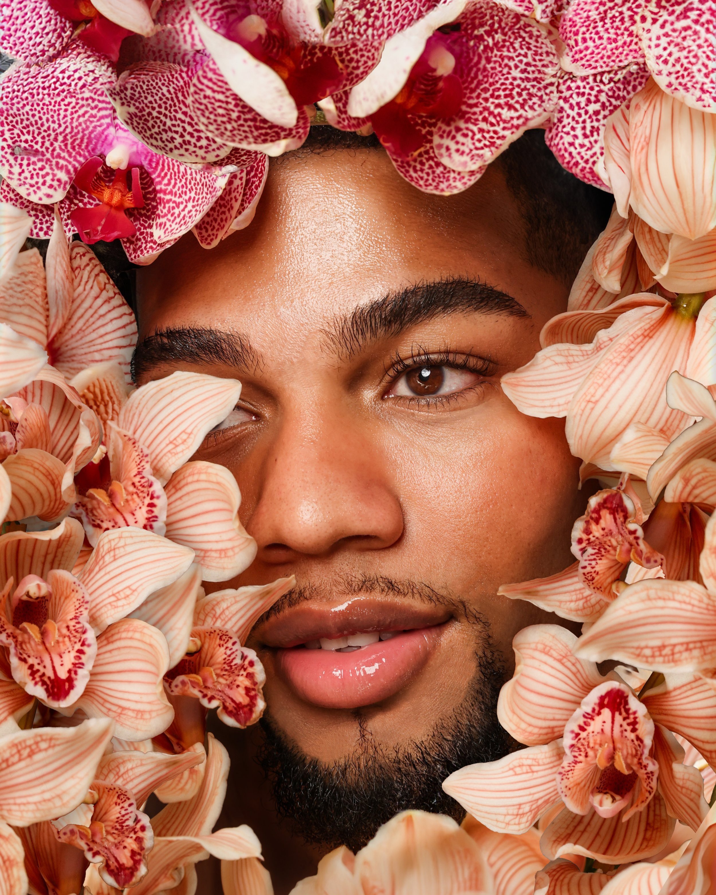 A close-up portrait of a young man with short dark hair and a beard, surrounded by pink and white orchids.