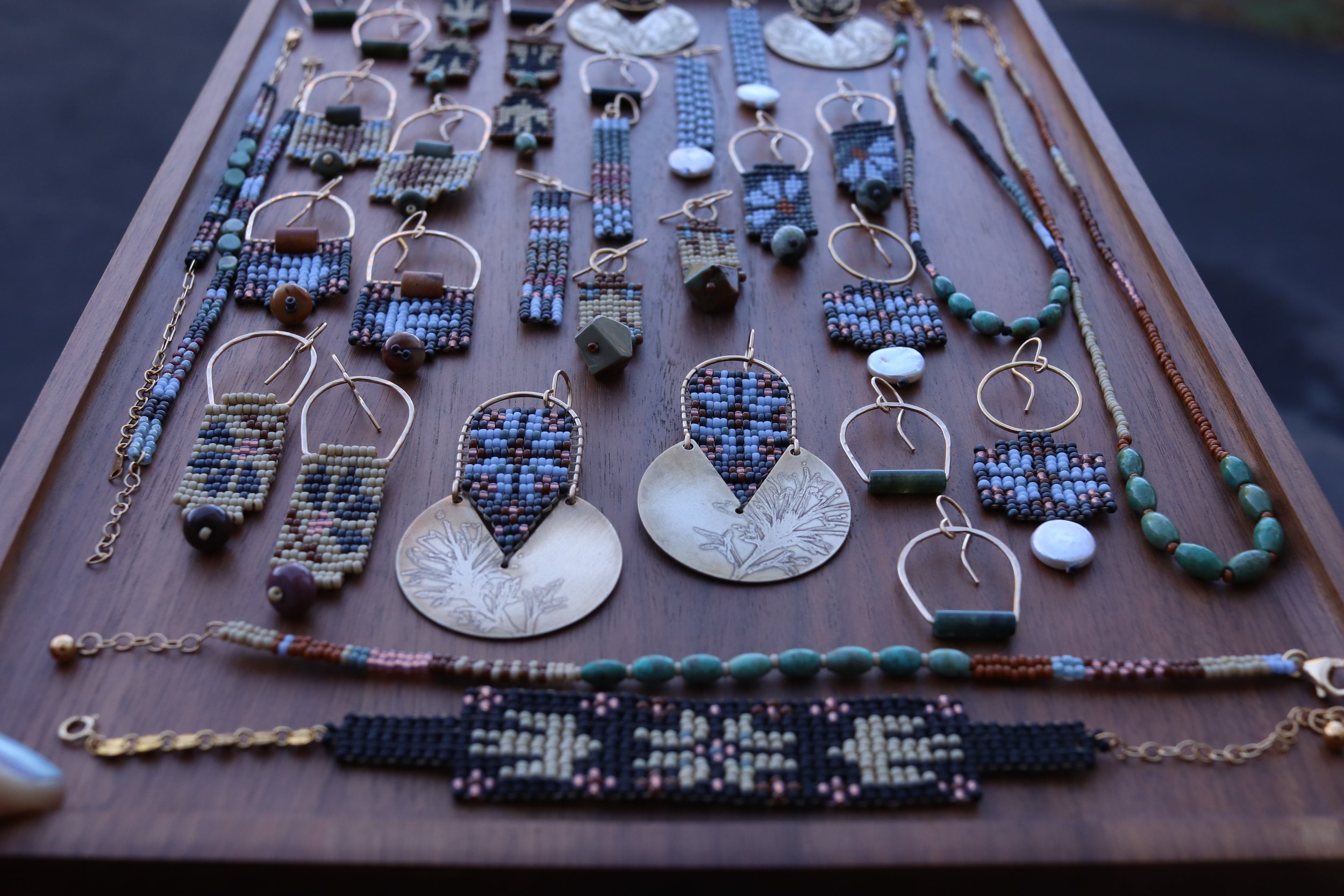 Collection of handmade beaded earrings and necklaces arranged on a wooden tray, with some decorative stones and dried plant at the bottom.