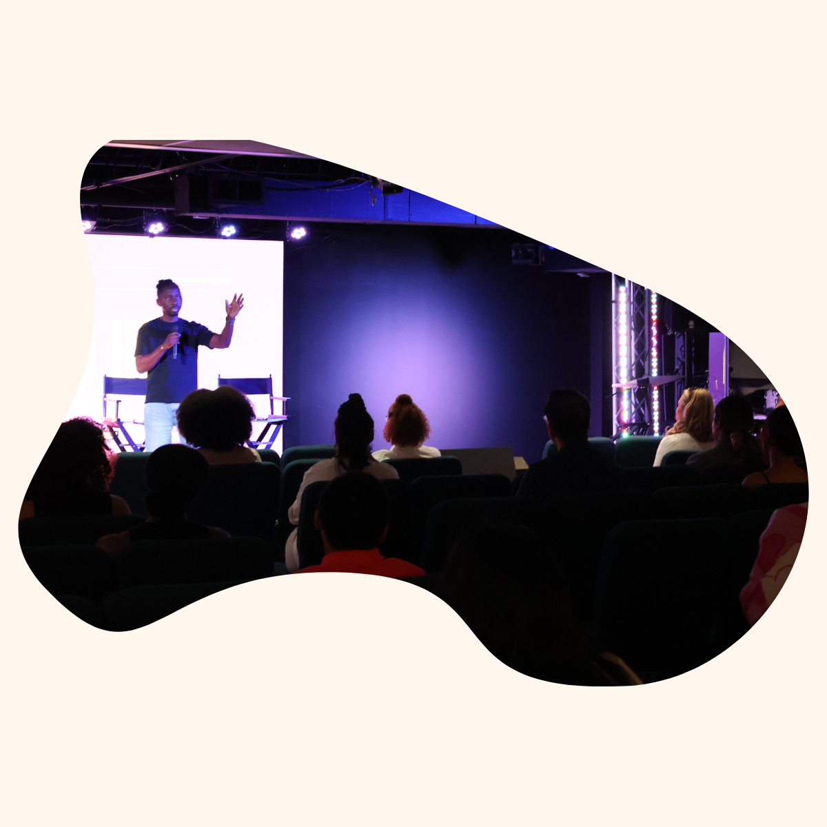 A person standing on stage giving a presentation to an audience in a darkened room with stage lighting.