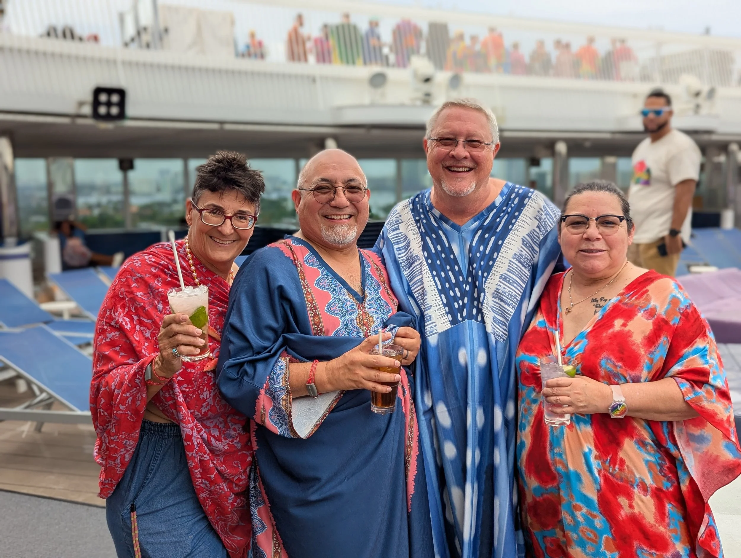 Four Big Fabulous Cruise Sailors from 2025 smiling on a cruise ship deck, wearing colorful clothing, holding drinks, with lounge chairs and a railing in the background.