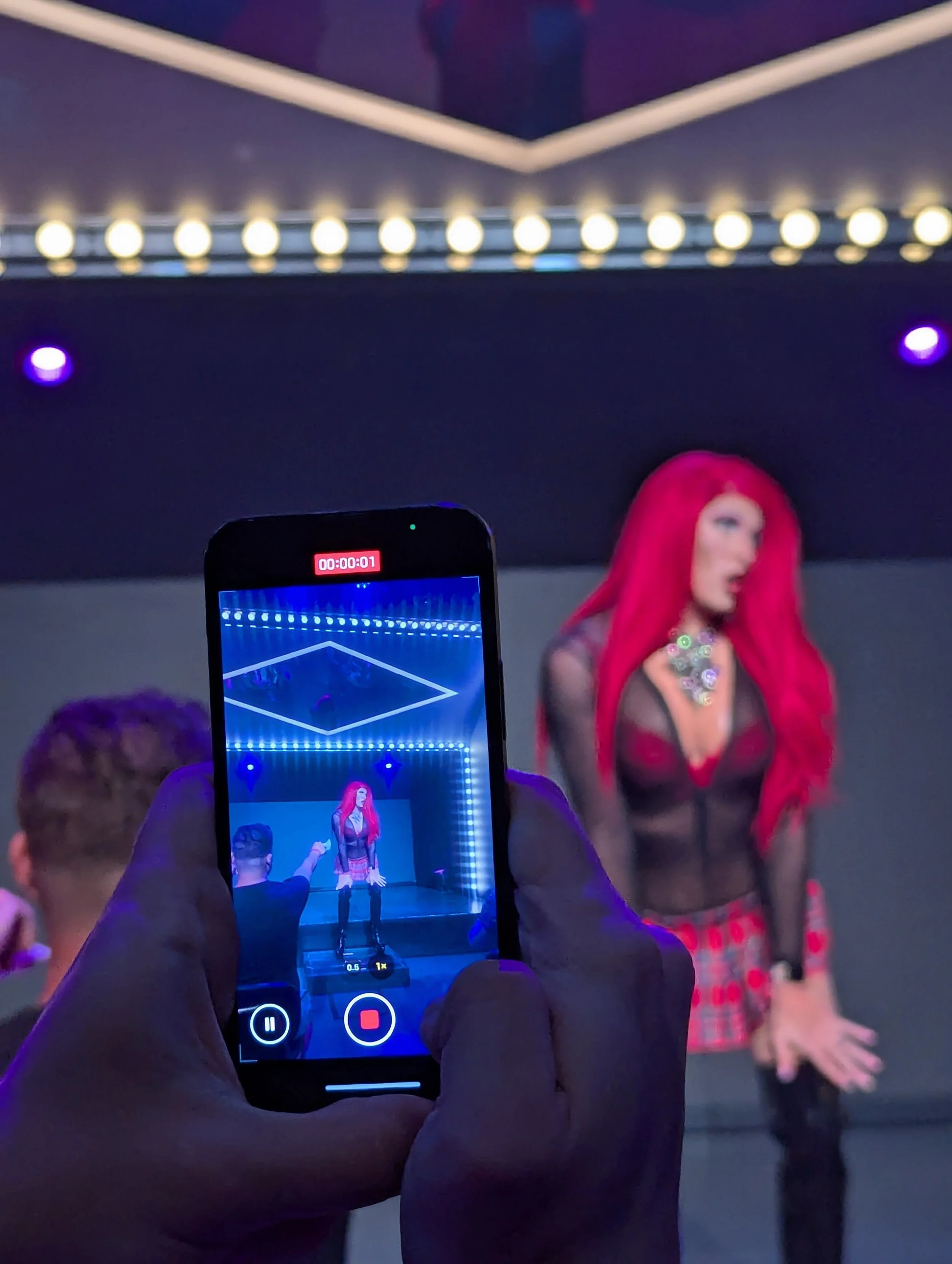 Person recording one of the Sisters in Action - Betty SiSonnet - with bright red hair on a smartphone during a performance or photoshoot in a studio with blue lighting and a dark background.