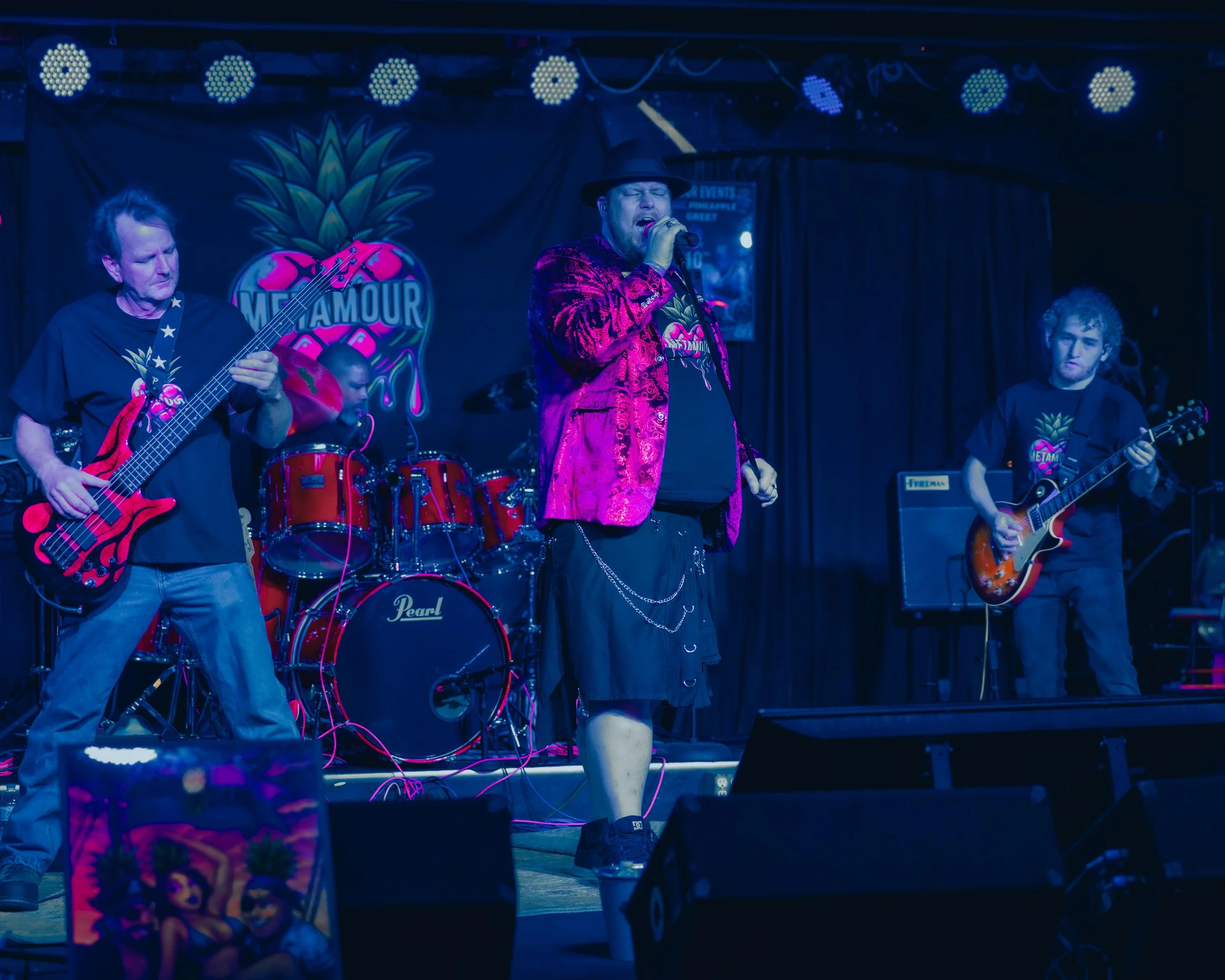 A band performing live on stage with four members, including a singer in a pink blazer and hat, a bassist, a drummer, and a guitarist, with stage lighting and a backdrop featuring a pineapple and the word 'MetalMOUR'.