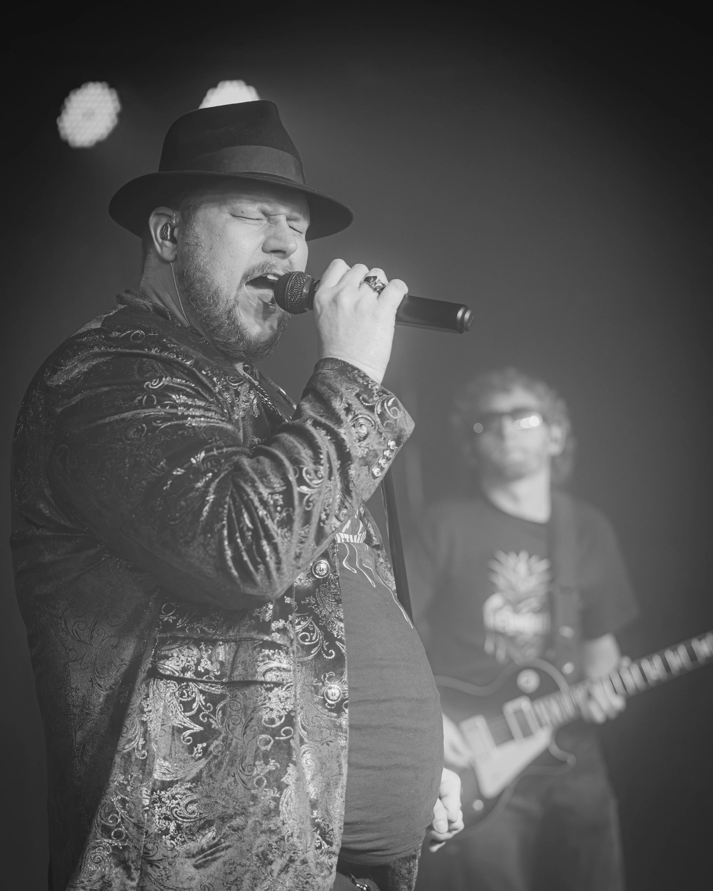 A man with a beard singing into a microphone, wearing a fedora hat and a detailed jacket, with another man in the background playing an electric guitar, both on stage in a black and white photo.