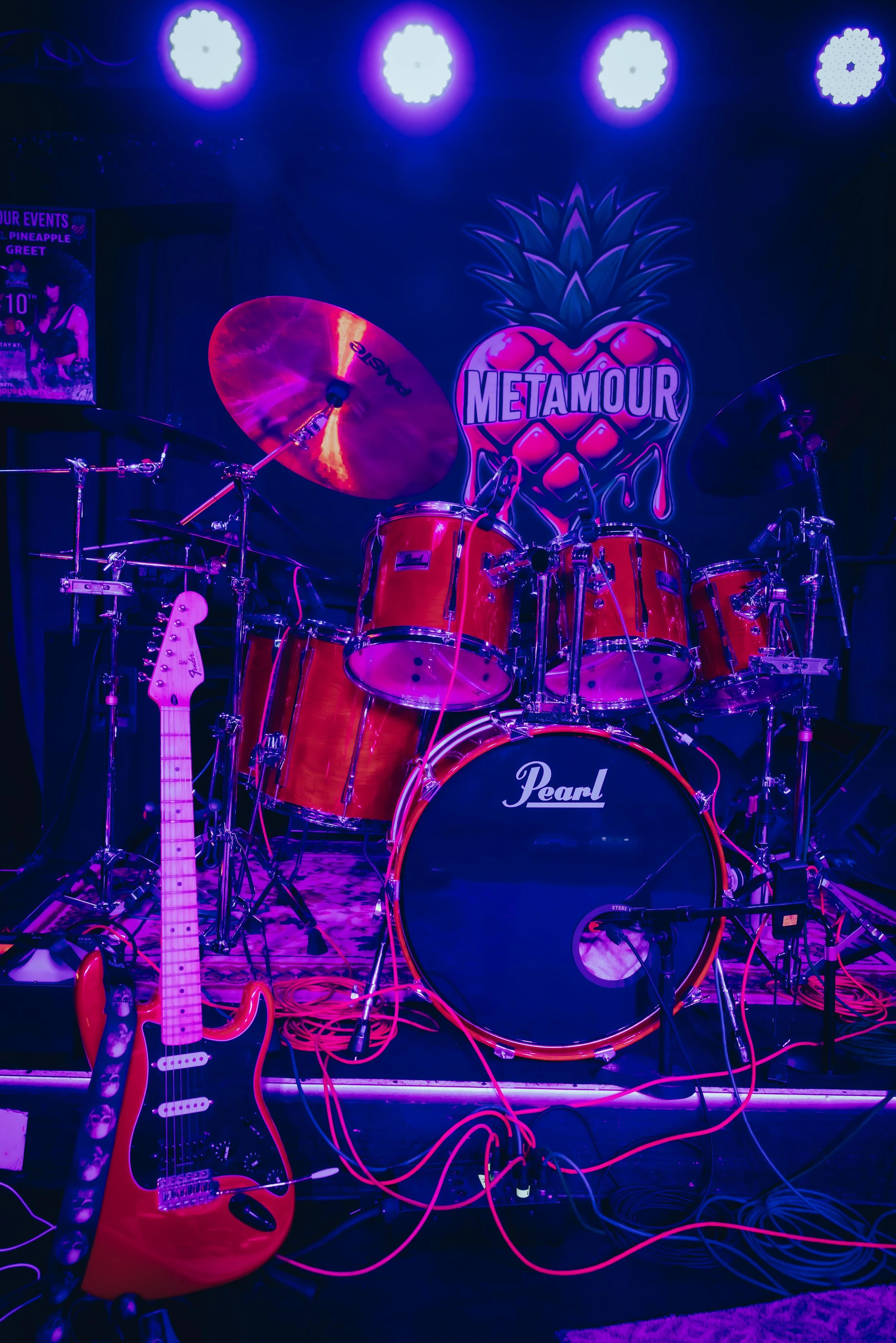 Stage with drum kit, electric guitar, and colorful artificial pineapple logo with the word 'METAMOUR,' illuminated by purple stage lighting.
