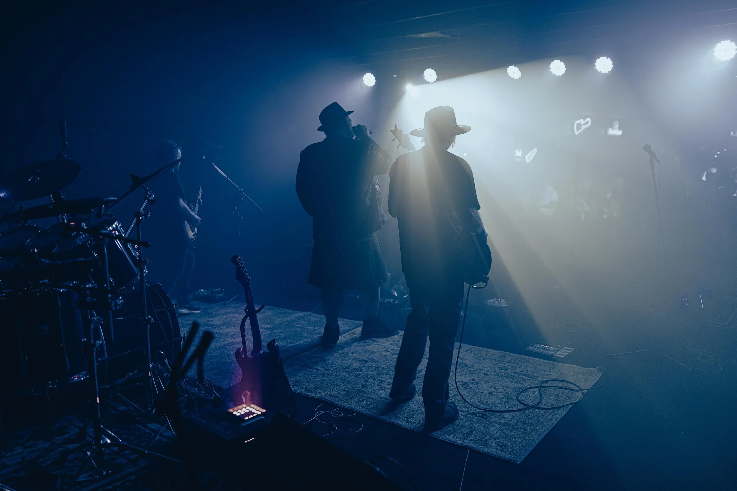 Musicians performing on stage under blue and white lights, with silhouettes of band members and musical instruments visible.