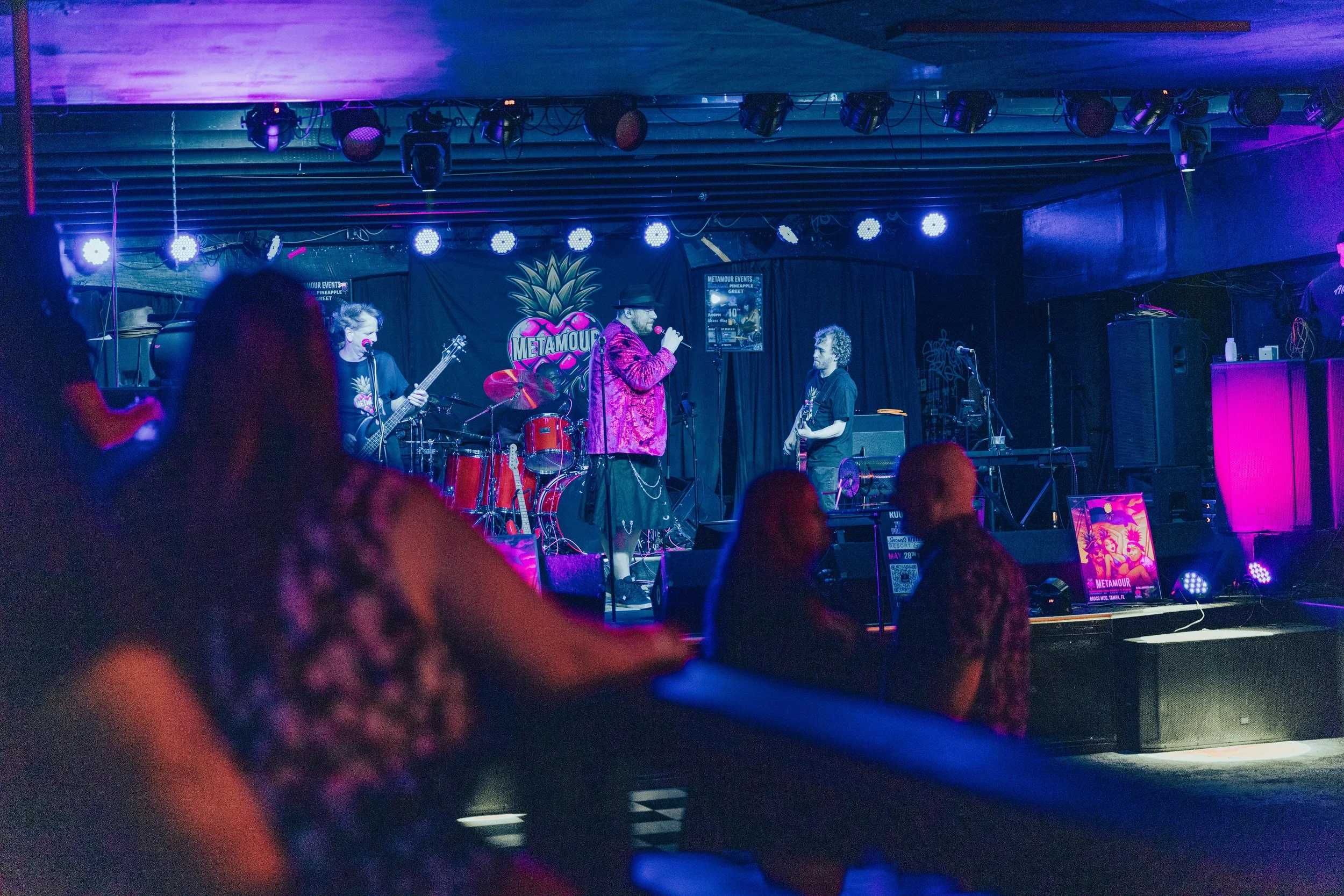 A live music performance on a small stage with a band and an audience. The stage has a drum set, guitars, and microphones, with colorful lighting and a Metamour sign in the background.