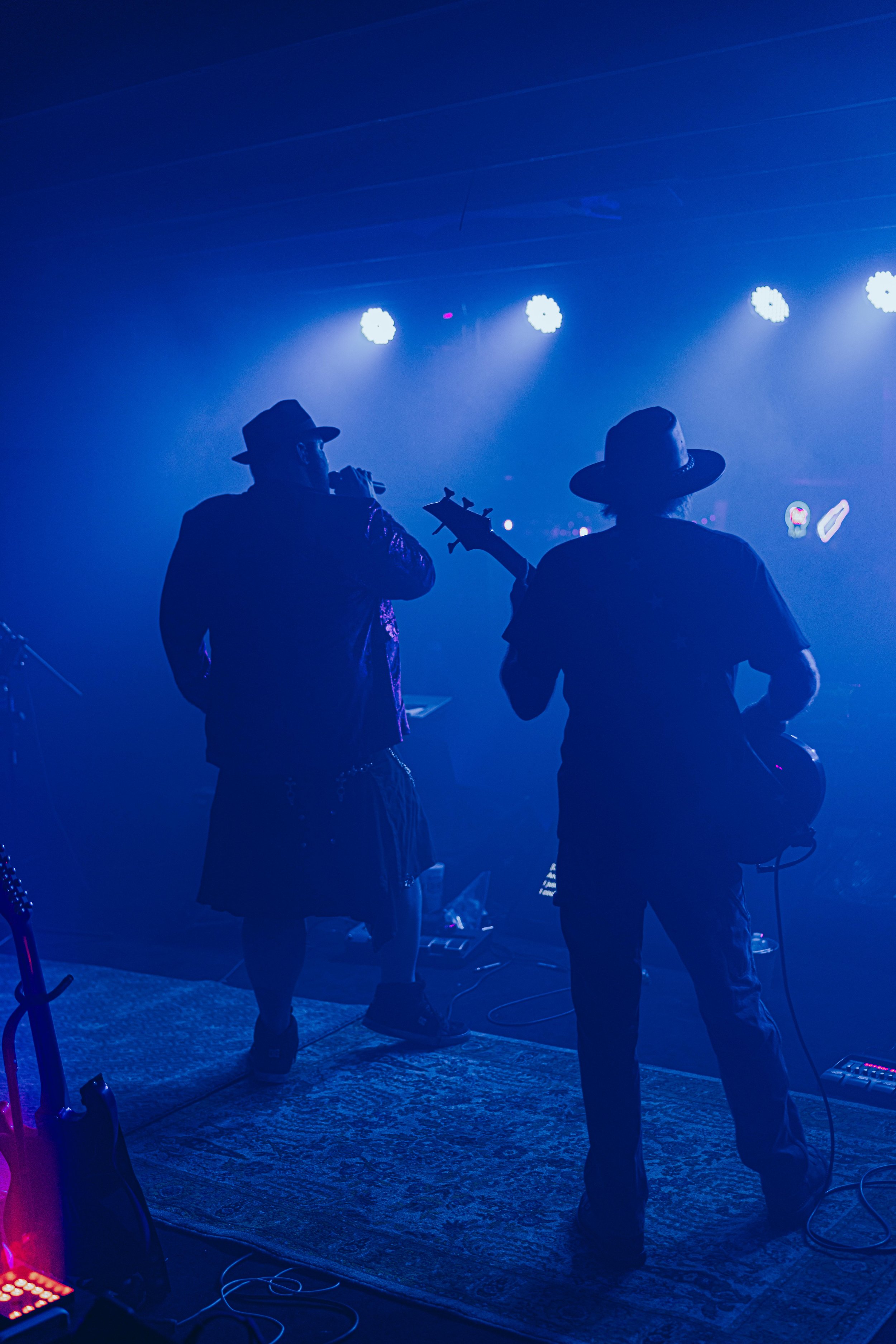 Two musicians on stage, one singing and the other playing guitar, under blue stage lights, both wearing hats.