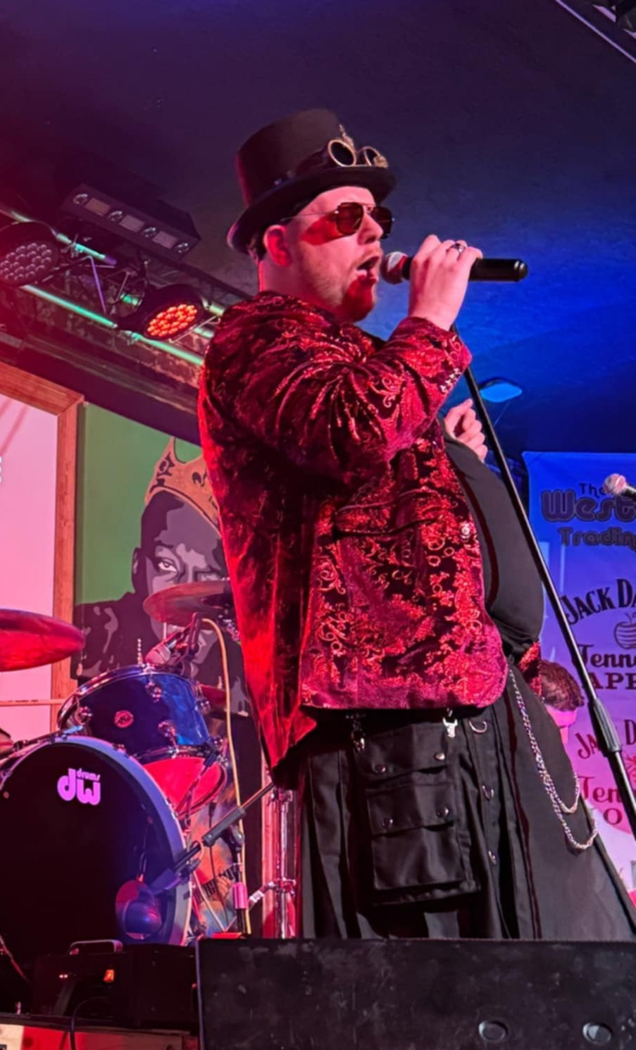 Musician in steampunk outfit performing on stage with microphone, wearing a red velvet jacket, top hat, and sunglasses, with drum set and colorful stage lights in the background.