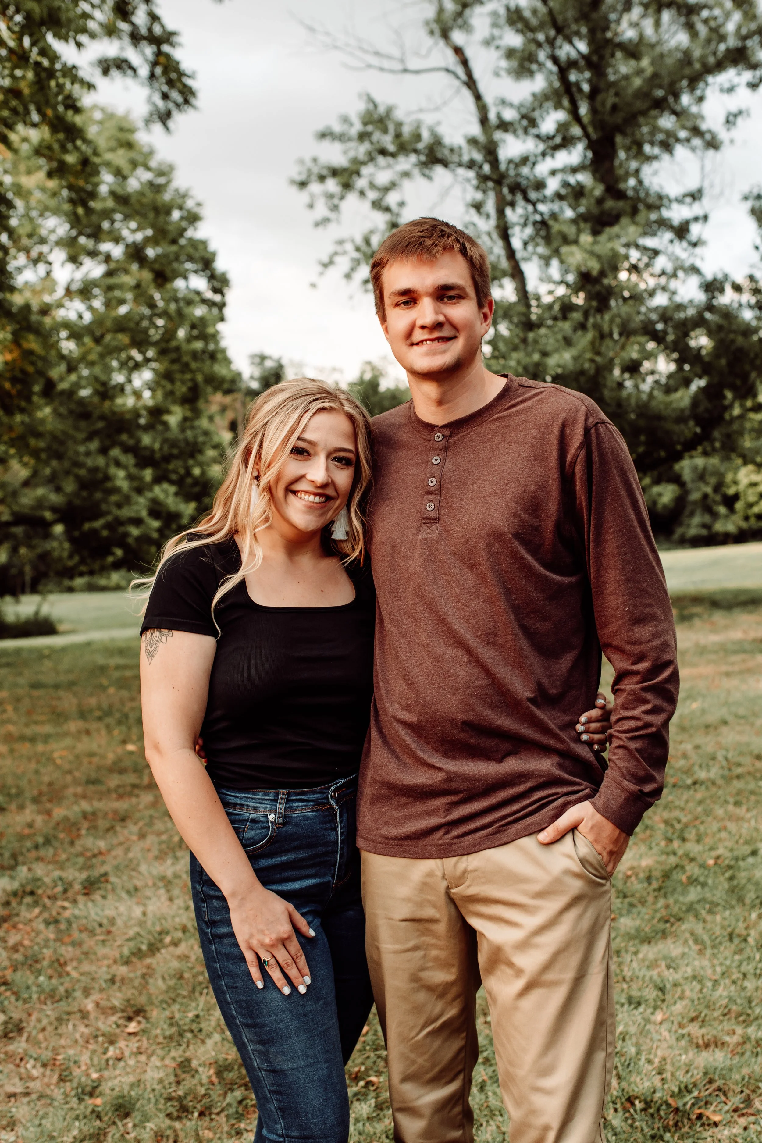 A young couple standing close together outdoors in a park with trees in the background. The woman has blonde hair, is wearing a black top and jeans, and is smiling. The man is taller, has brown hair, is wearing a brown long-sleeve shirt and beige pants, and is also smiling.