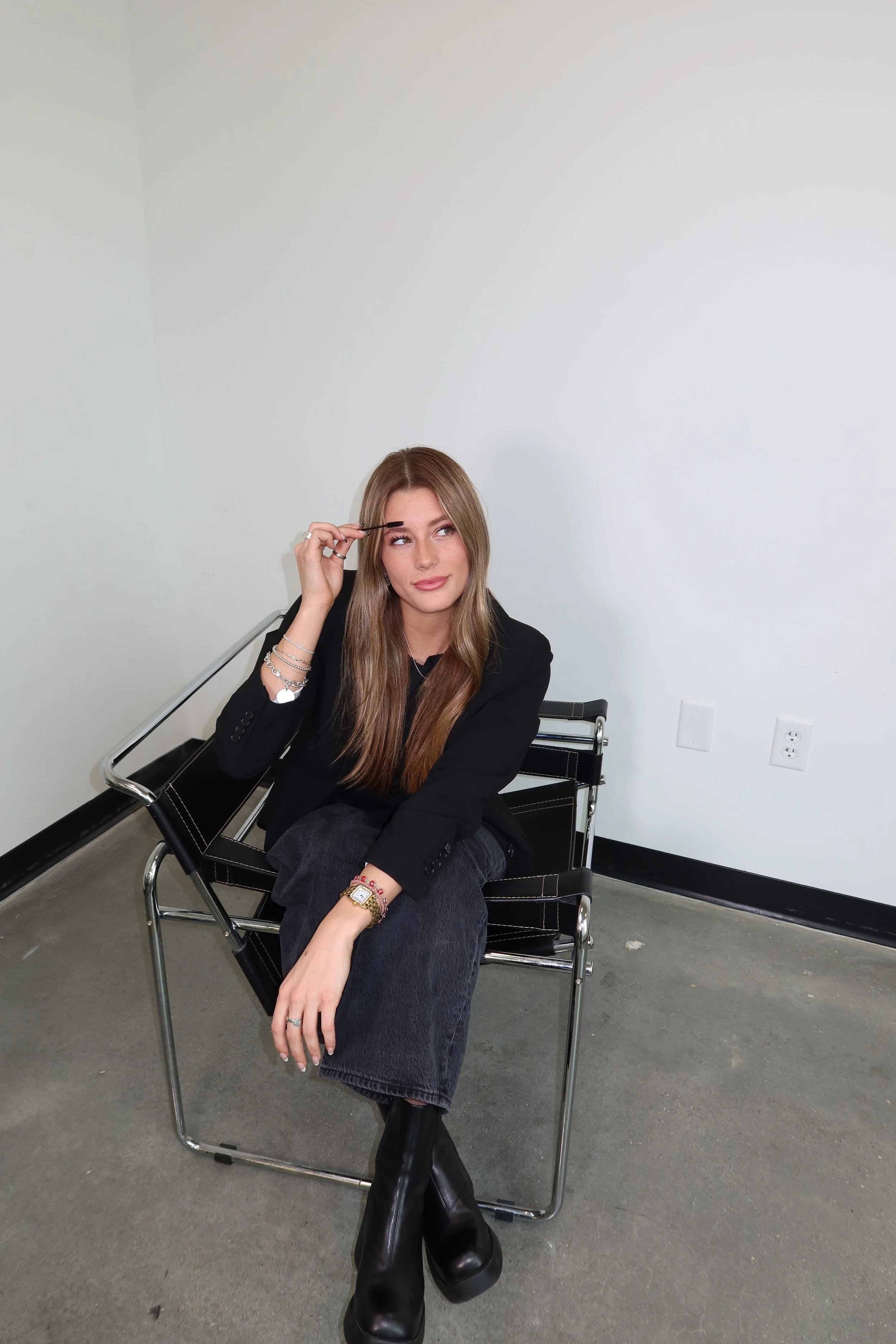 A young woman with long light brown hair sits on a black chair in a minimalist room, wearing a black blazer, dark jeans, and black platform boots, holding a makeup brush to her eyebrows.