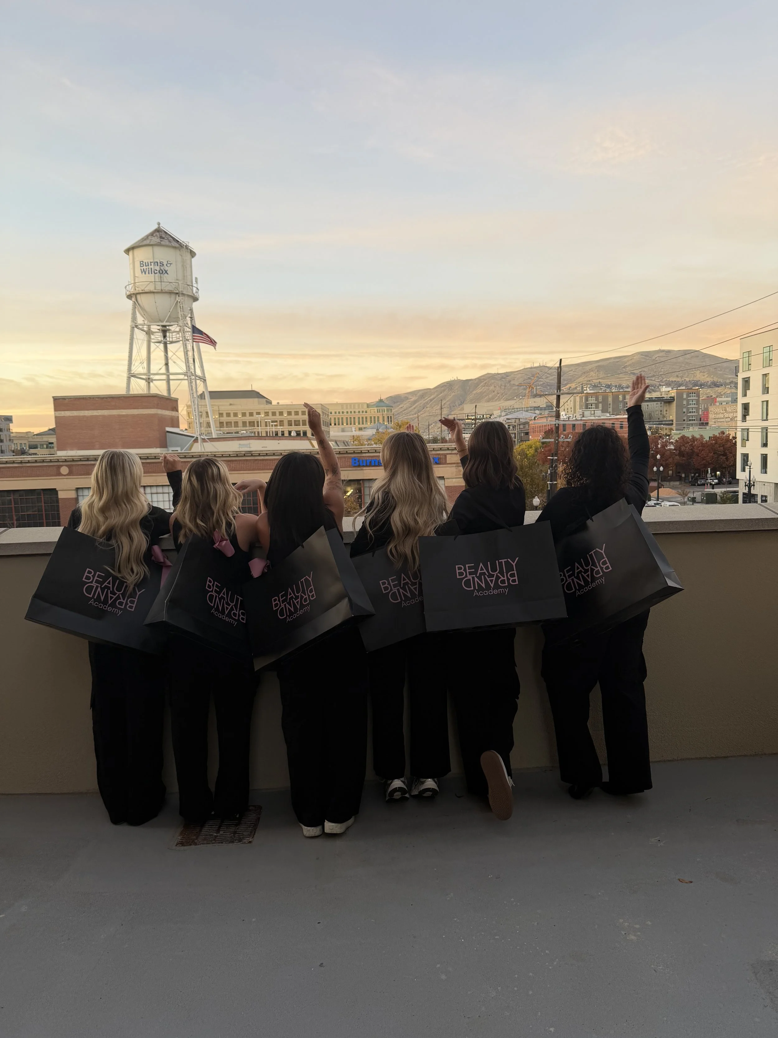 Five women from behind, holding large shopping bags with 'Beauty Brand Academy' written on them, overlooking a cityscape at sunset, with buildings, a water tower, and mountains in the background.