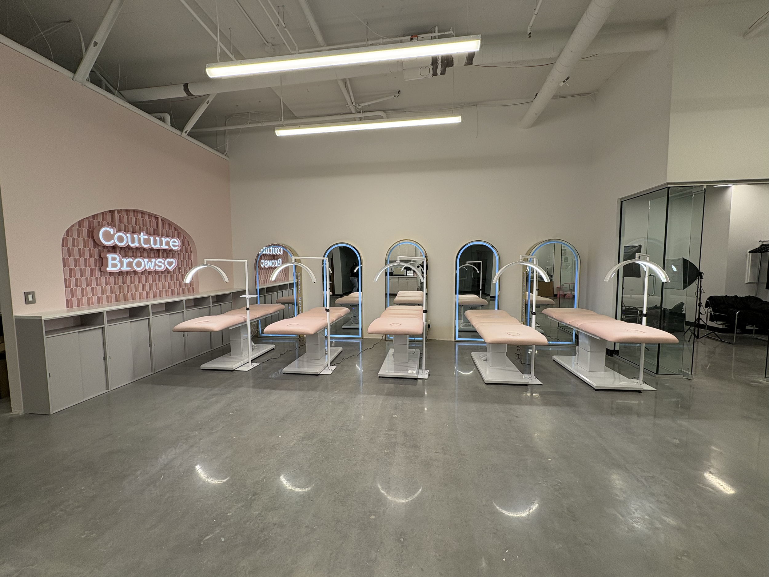 A modern beauty salon room with seven pink treatment beds and individual lamps, mirrors with arched frames, and a neon sign that reads 'Couture Brows' on a pink wall.