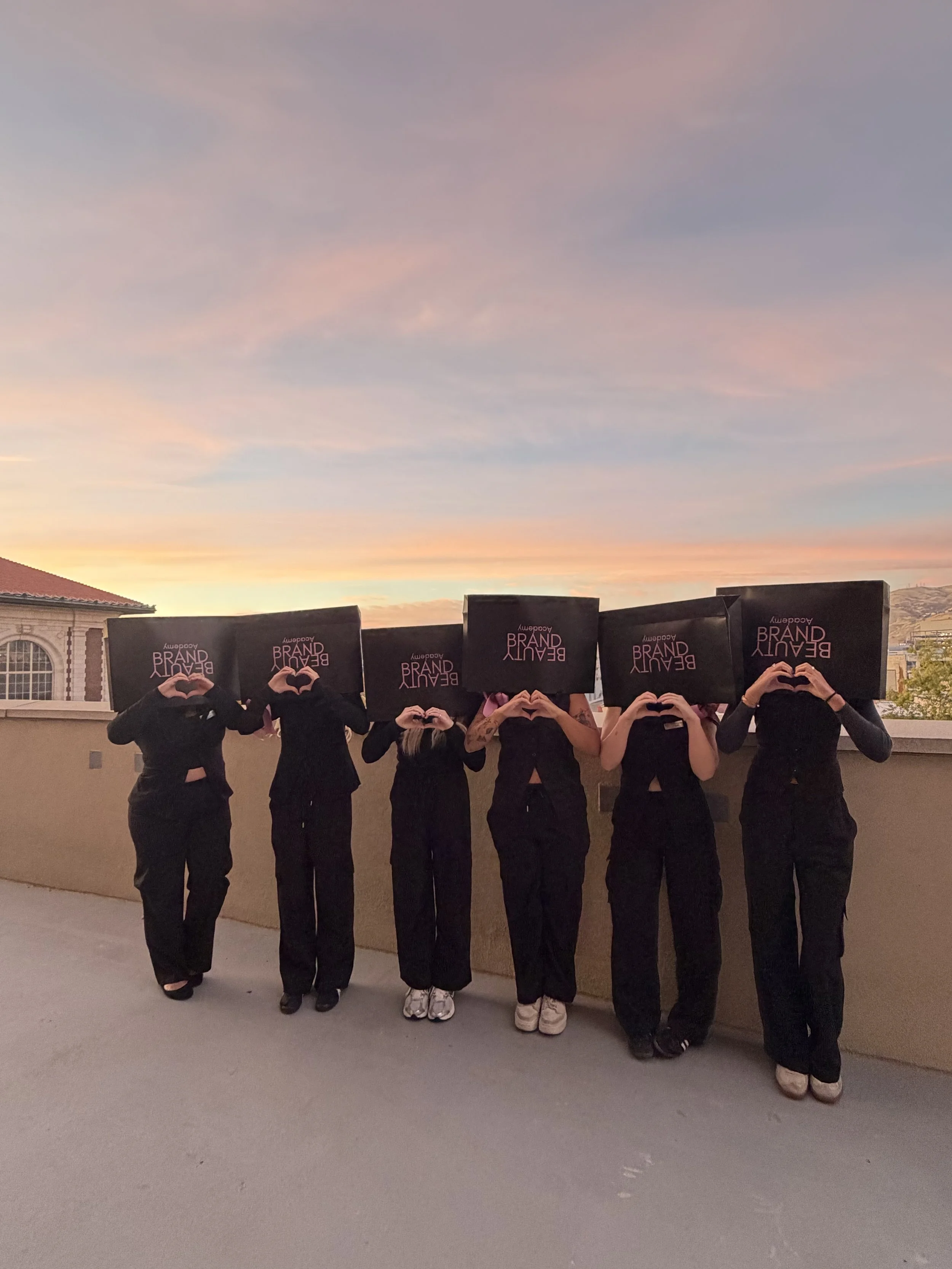 Six people dressed in black holding large black boxes on their heads, forming heart shapes with their hands. The boxes have the words 'Beauty Brand' printed on them. The scene is outdoors during sunset with a pink and blue sky.