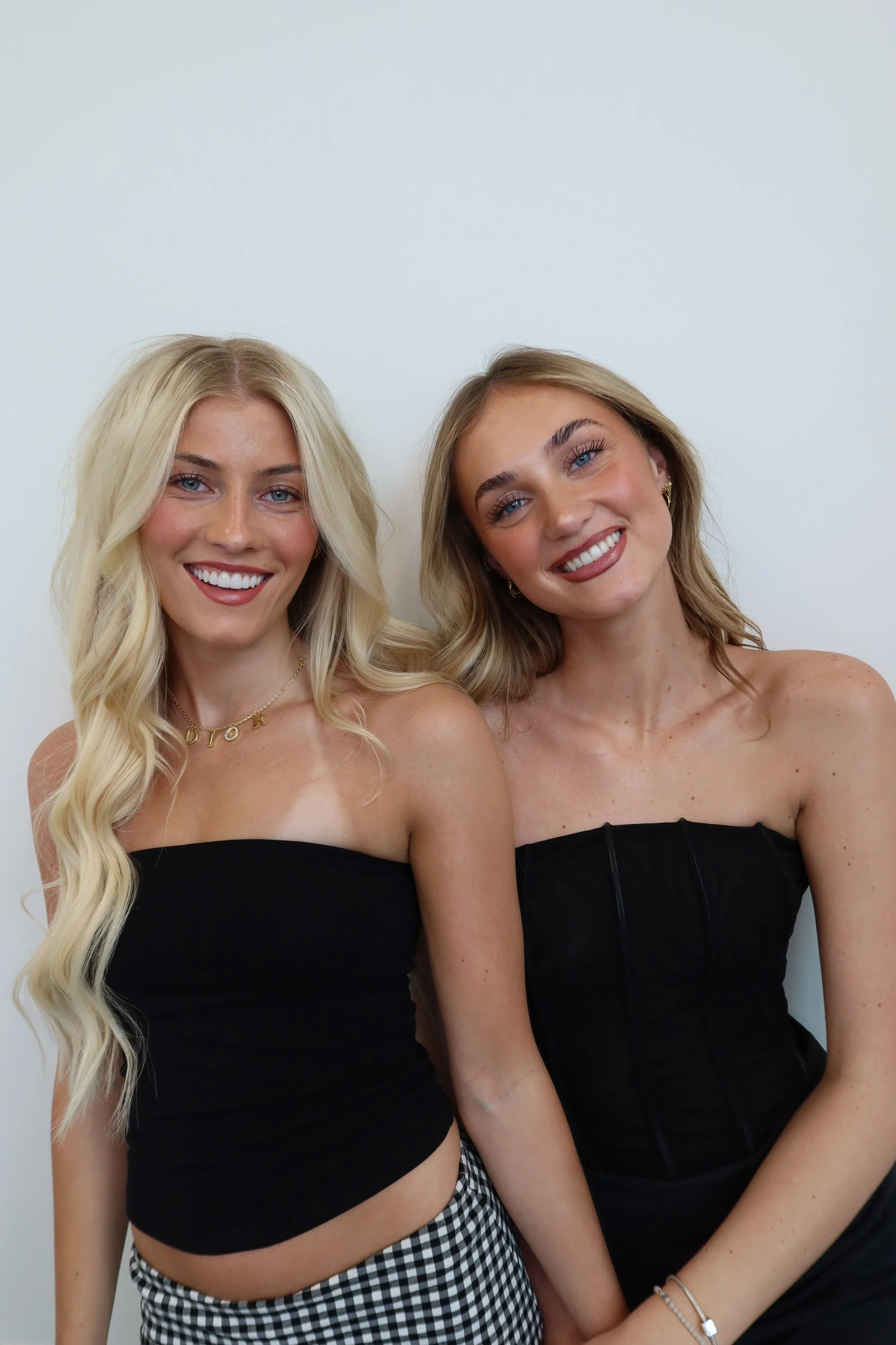 Two young women with blonde hair smiling, standing close together in front of a plain white wall. Both are wearing strapless black tops, and the woman on the left has wavy hair and checkered pants while the woman on the right has straight hair.