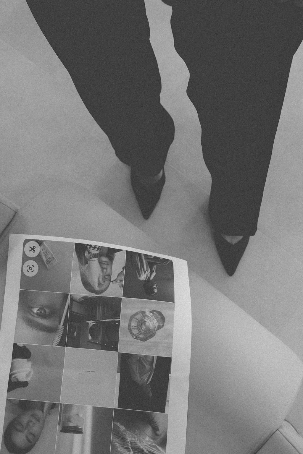 A person's legs and feet wearing black pointed-toe shoes and dark pants, standing on a light floor, next to a printed collage of photographs.
