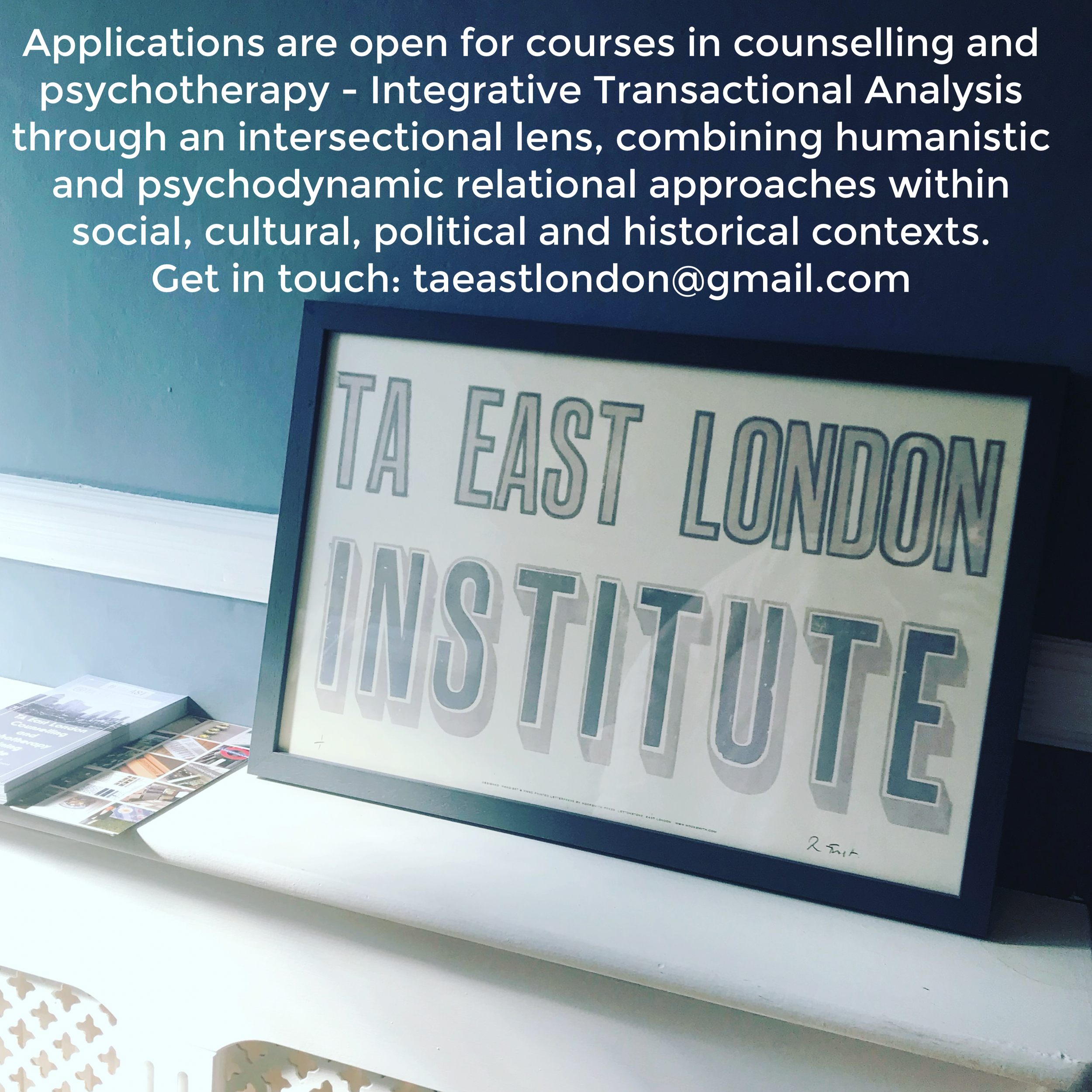 A framed sign on a table reads 'EAST LONDON INSTITUTE' with the words in bold, vintage-style lettering. To the left of the frame, there are brochures or pamphlets stacked on the table against a blue wall with a white baseboard.