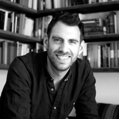 A black and white portrait of a smiling man with dark hair and a beard, wearing a dark shirt, sitting in front of a bookshelf filled with books.