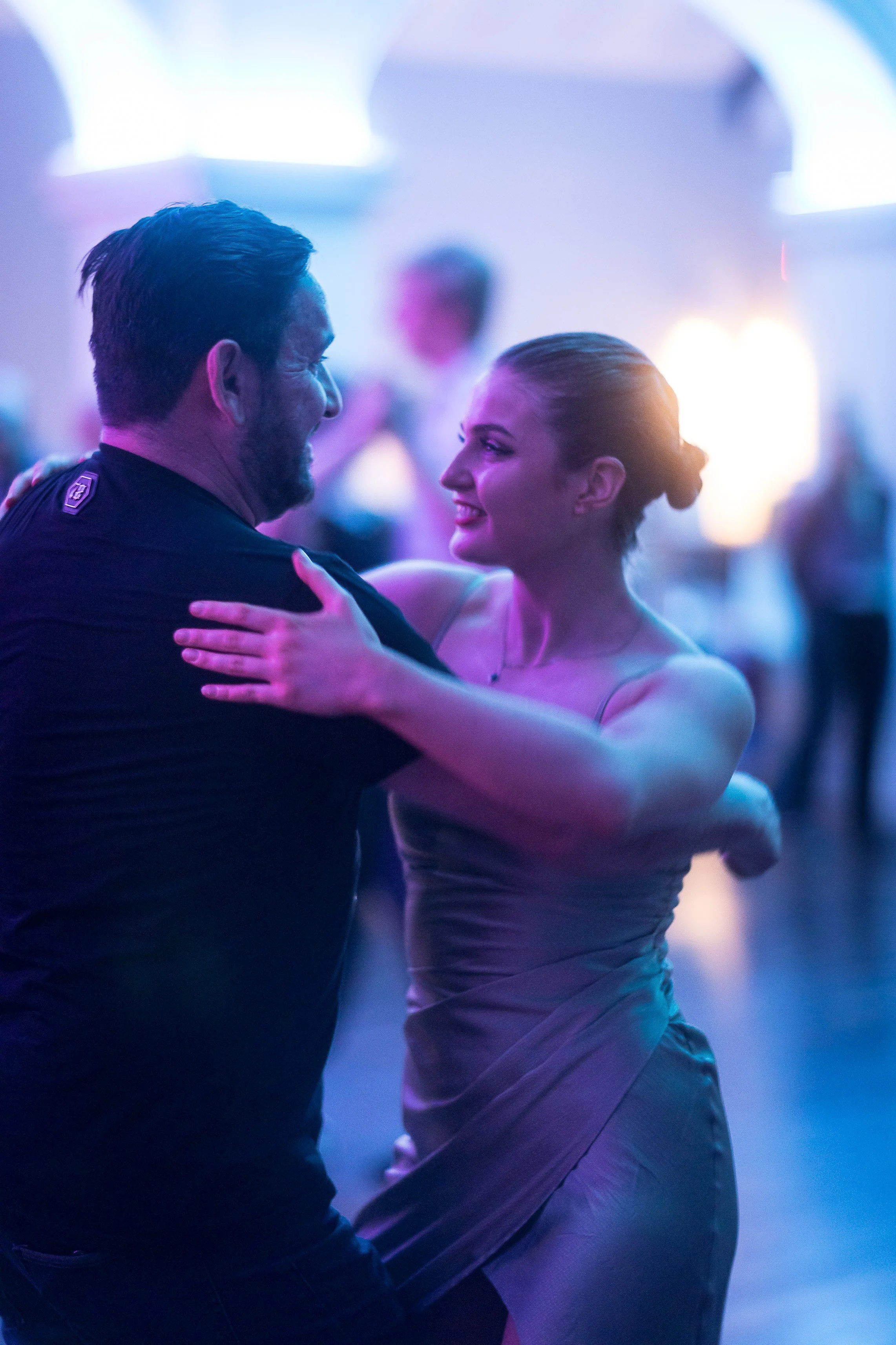 A man and woman dancing together at a party or event, with the woman smiling and looking into the man's eyes. The background is filled with colorful lighting.